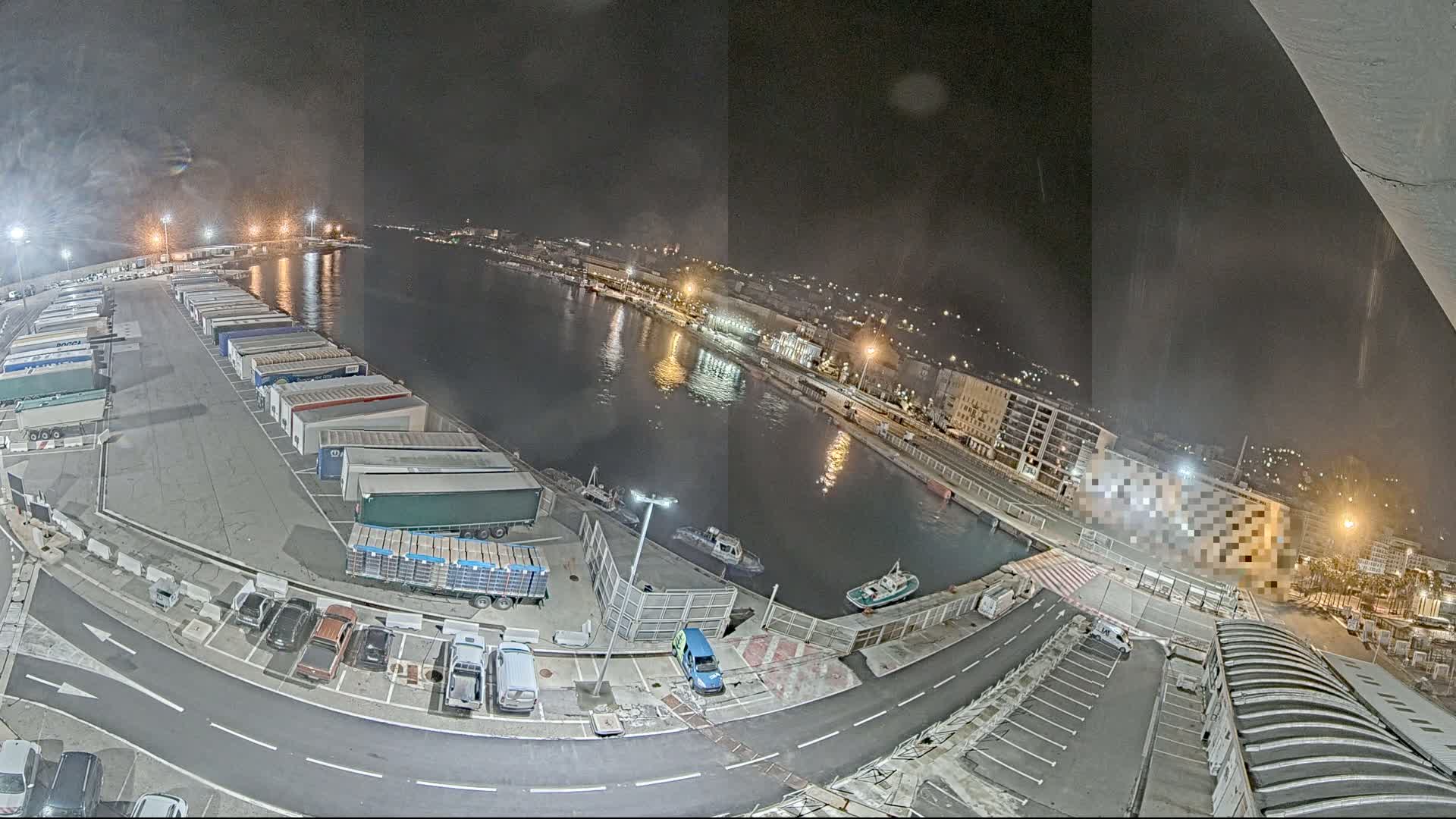 A nighttime aerial view of a harbor shows numerous trucks parked in a lot next to a calm body of water, with a few small boats and a city in the background.