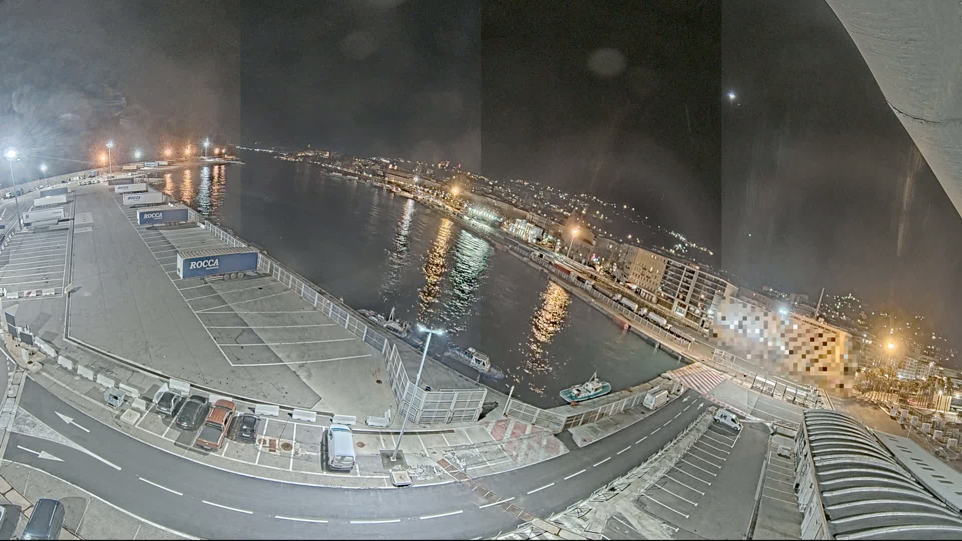 A nighttime high-angle view of a harbor with several parked trucks and cars, boats on the water, and city lights in the background.