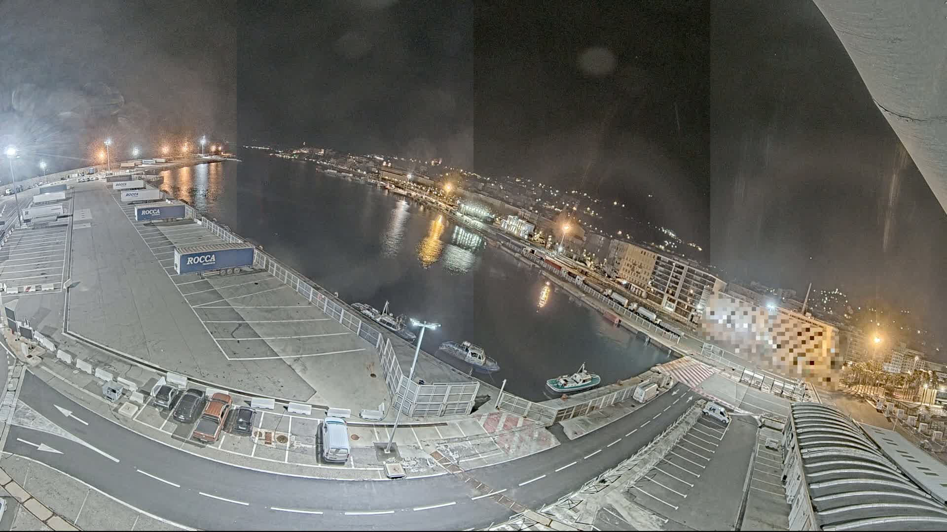 A nighttime aerial view of a harbor shows a mostly empty parking lot with several trucks and cars, a calm body of water with small boats, and a city with lit buildings in the background.