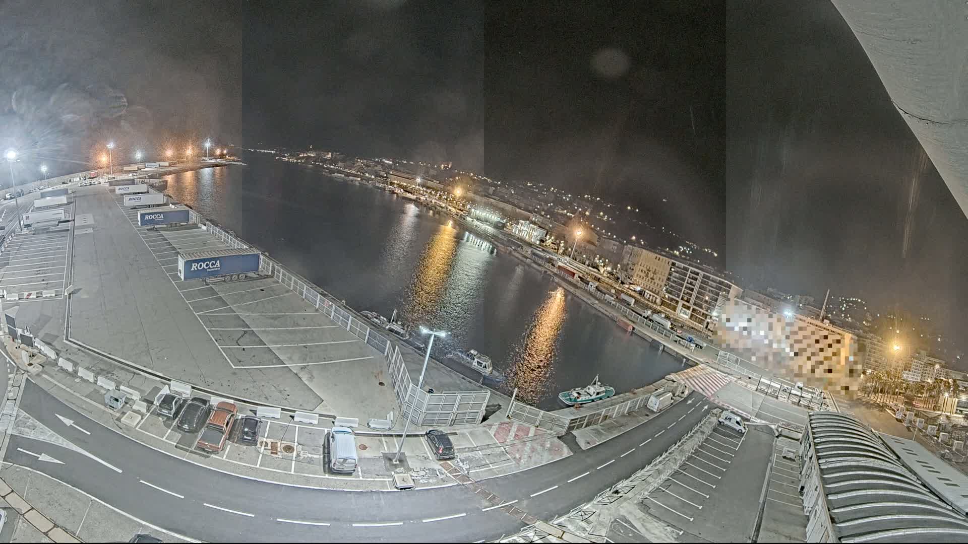 A nighttime aerial view of a harbor, with a parking lot filled with semi-trucks and cars, and a calm body of water reflecting city lights.