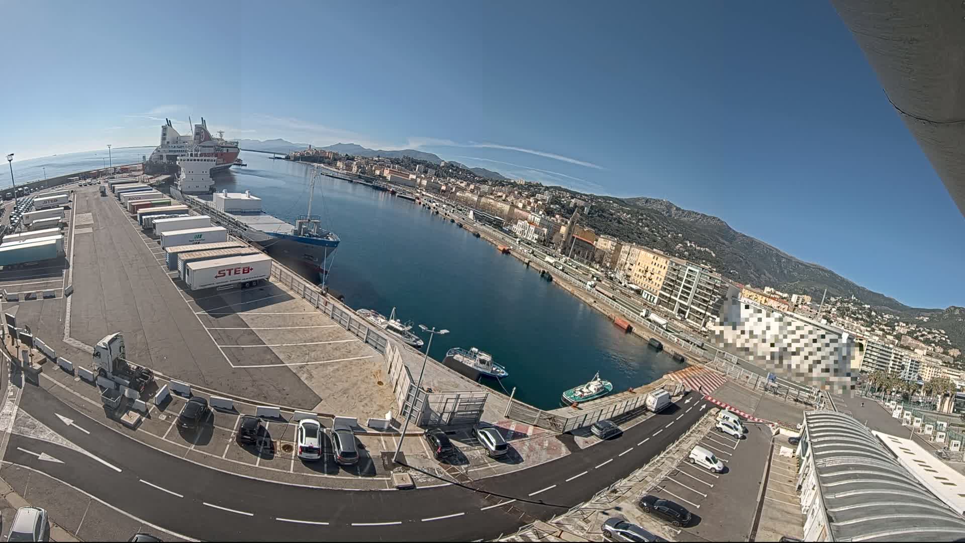 A harbor on a sunny day with several ships docked, numerous semi-trailer trucks parked in a lot, cars in a parking lot, and buildings lining the waterfront.