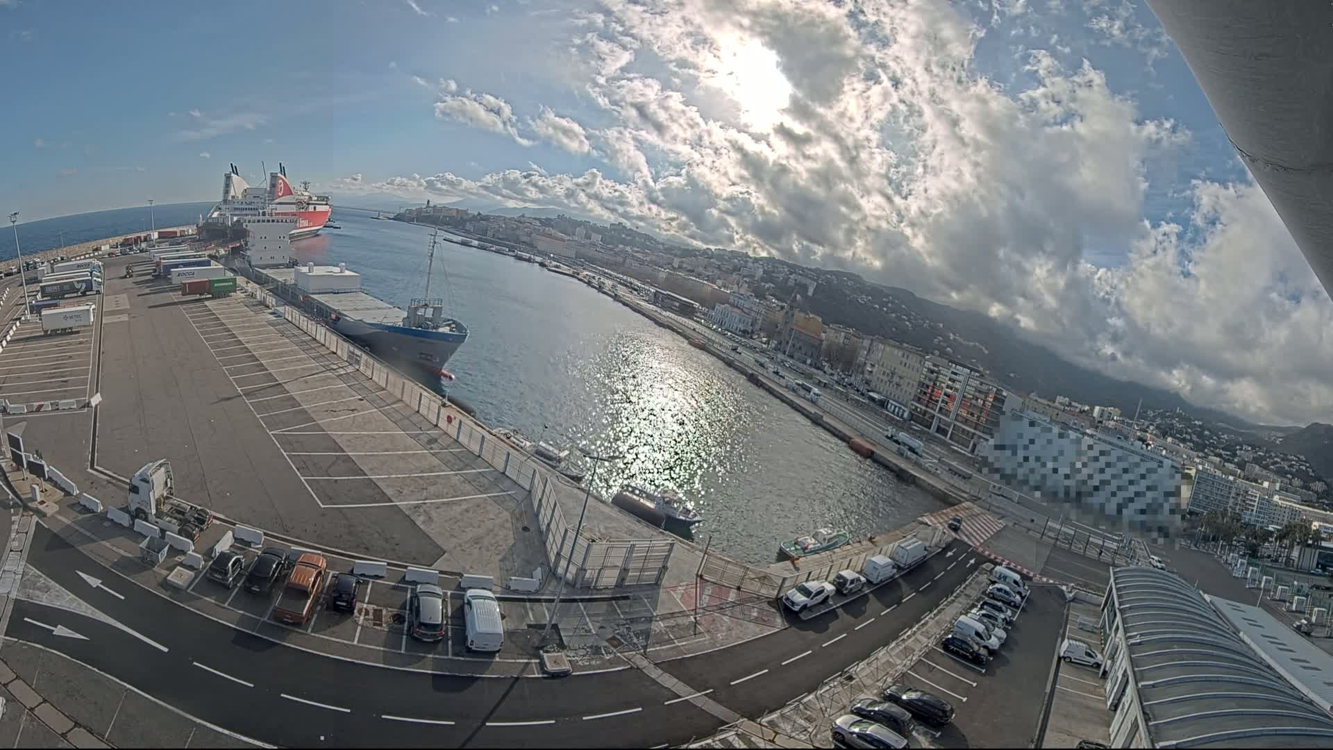 A sunny day with partly cloudy skies shows a harbor with docked cargo ships and ferries, adjacent parking lots filled with cars and trucks, and a city in the background.