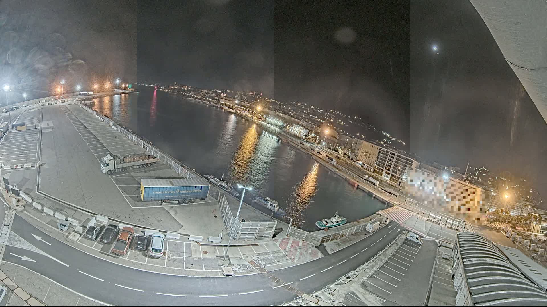 A nighttime aerial view of a harbor, showing a calm body of water reflecting city lights, alongside a parking lot with several vehicles and trucks.