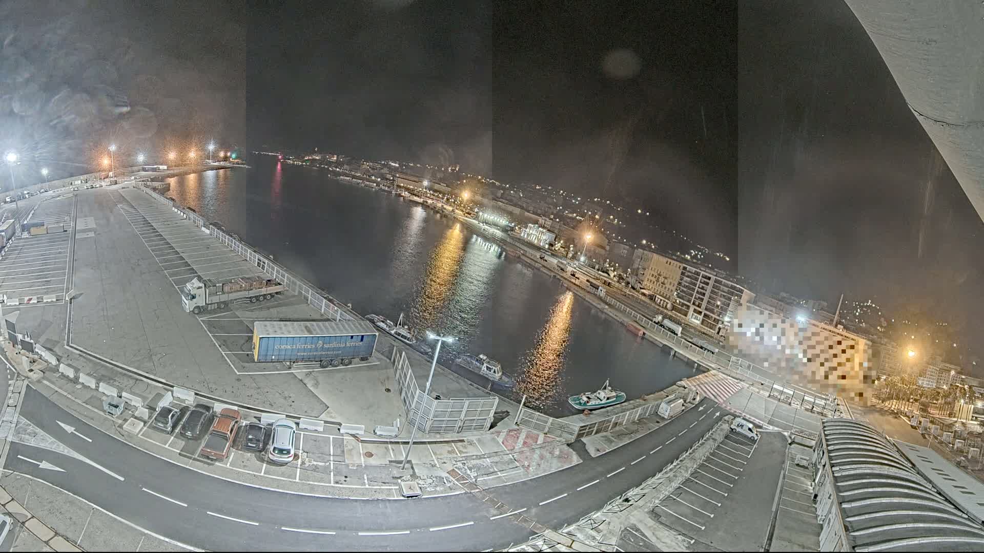 A nighttime aerial view of a harbor with a parking lot, several vehicles, and boats on the water.