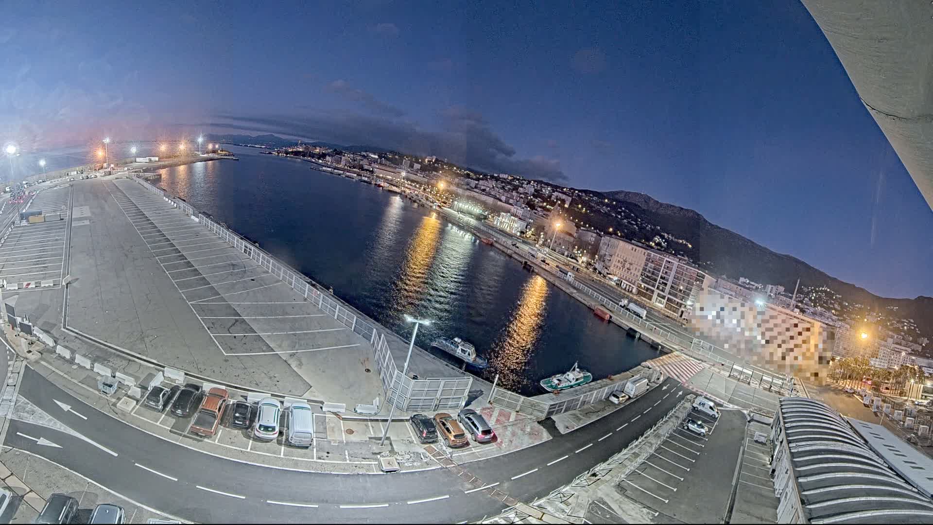 A mostly empty parking lot overlooks a calm harbor at twilight, with a city and mountains visible in the distance.