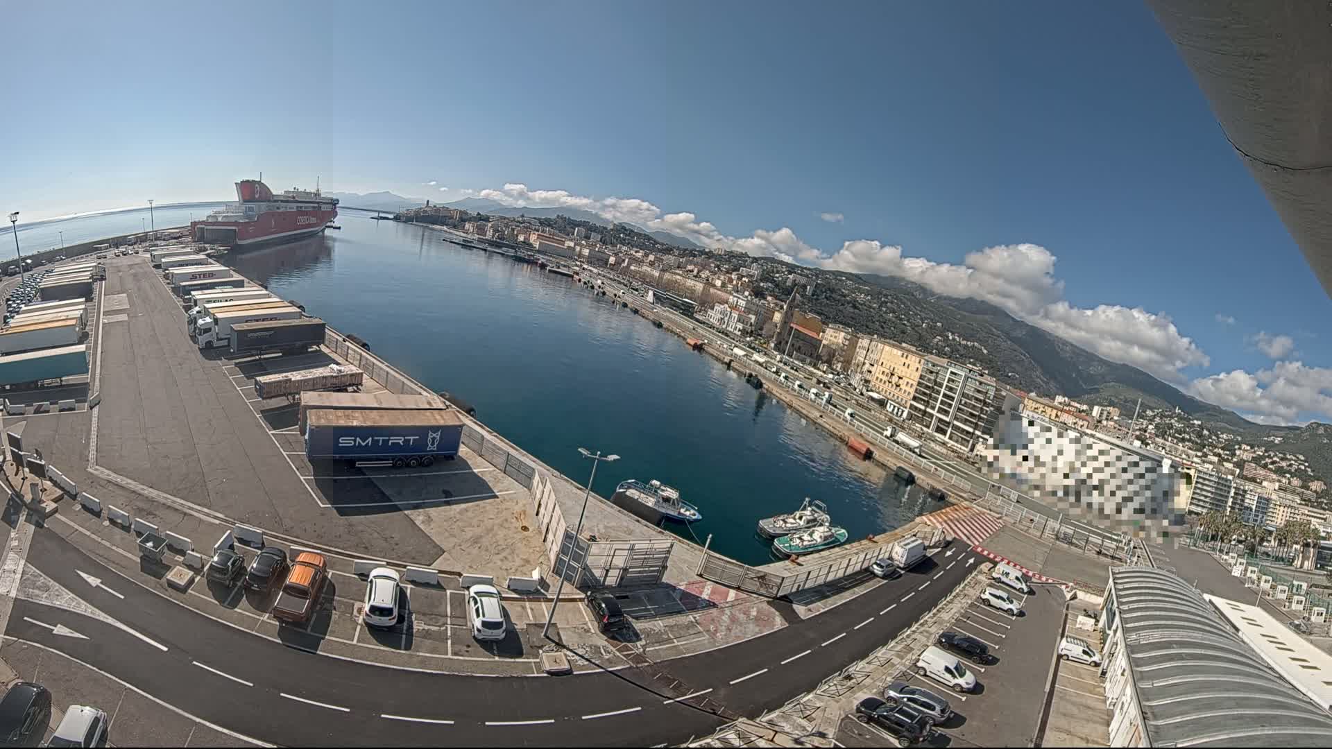 A large ferry is docked at a harbor, with numerous trucks and cars parked in the surrounding area, under a mostly sunny sky with some scattered clouds and mountains visible in the distance.