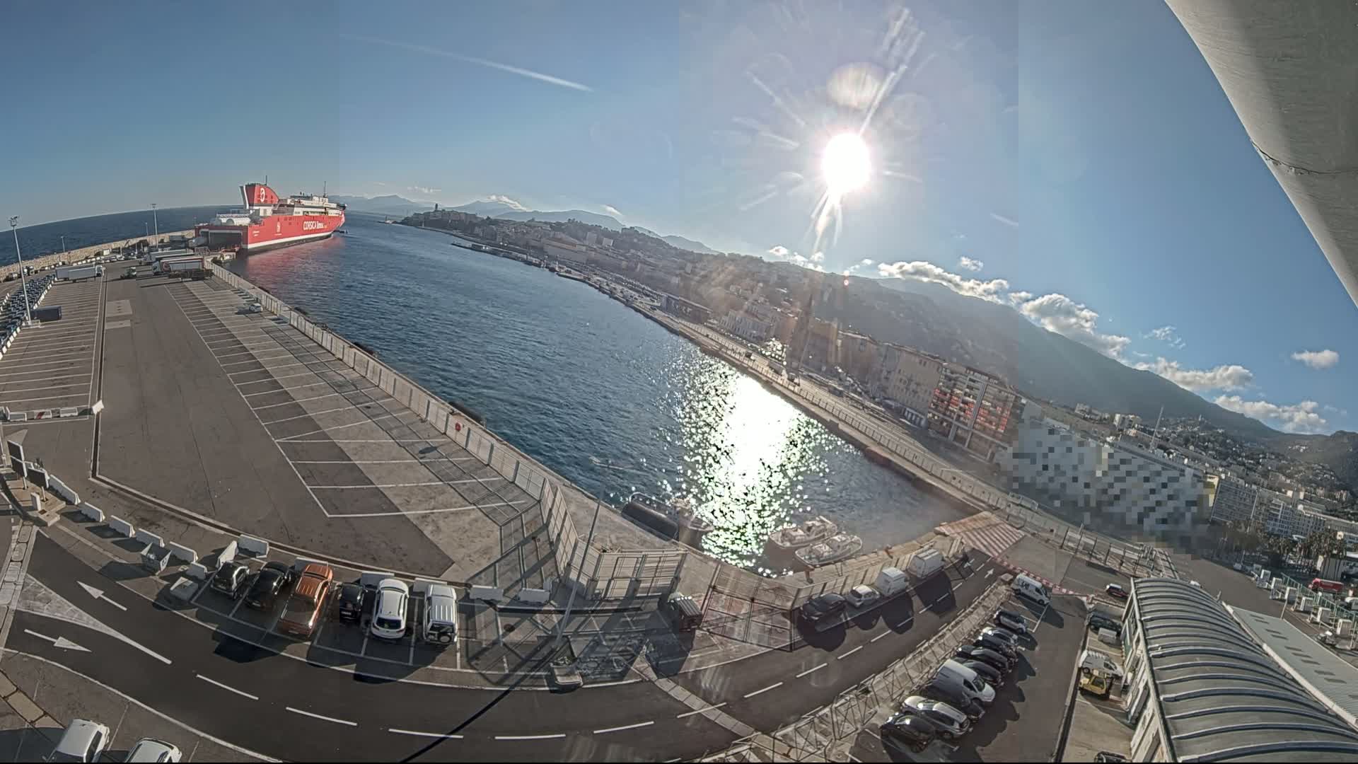 A large red ferry is docked at a harbor on a sunny day, with numerous vehicles parked in a lot nearby and a city and mountains visible in the background.