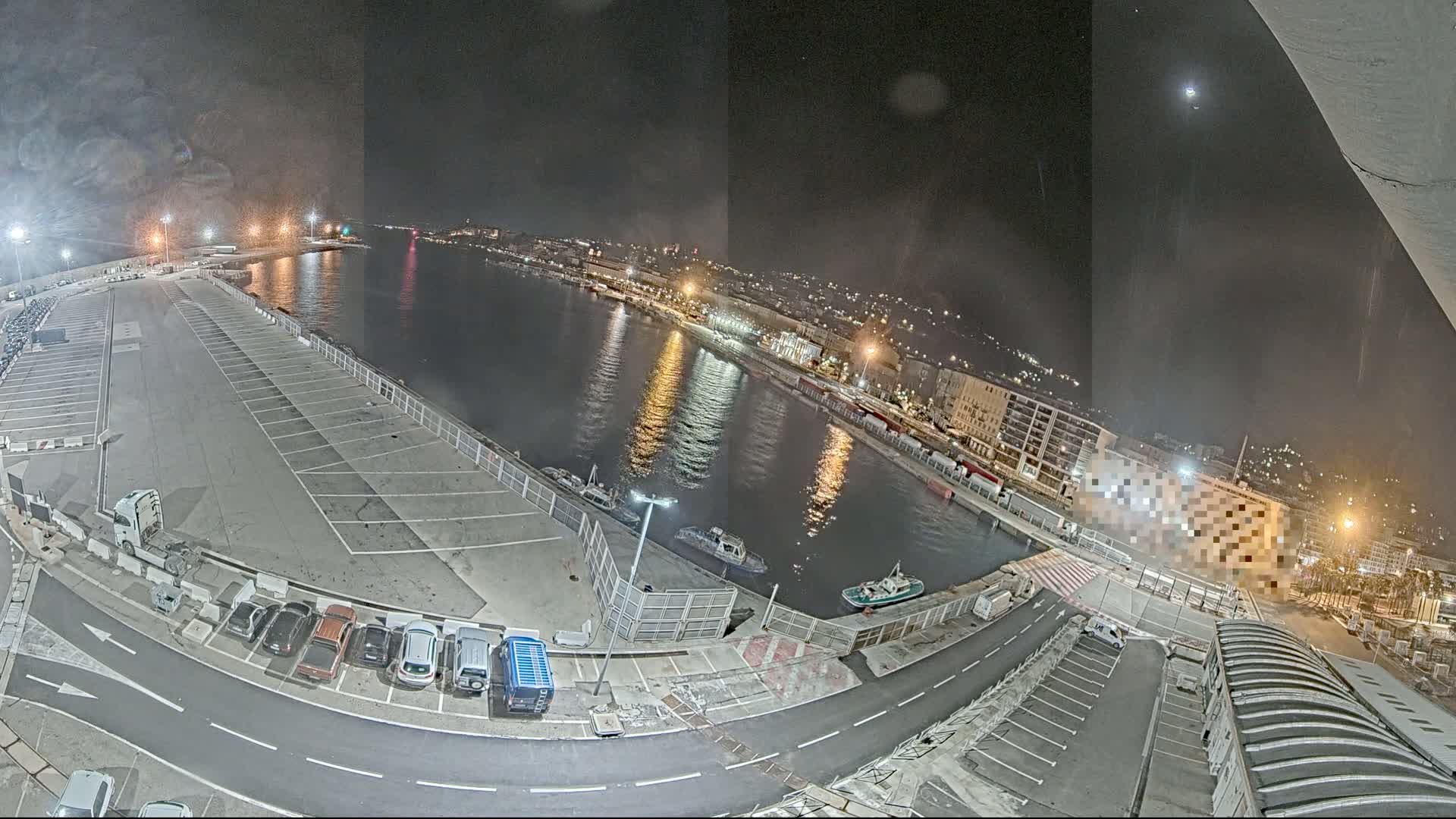 A nighttime aerial view of a harbor with a large empty parking lot, several small boats on the water, and city lights visible in the distance under a clear sky.