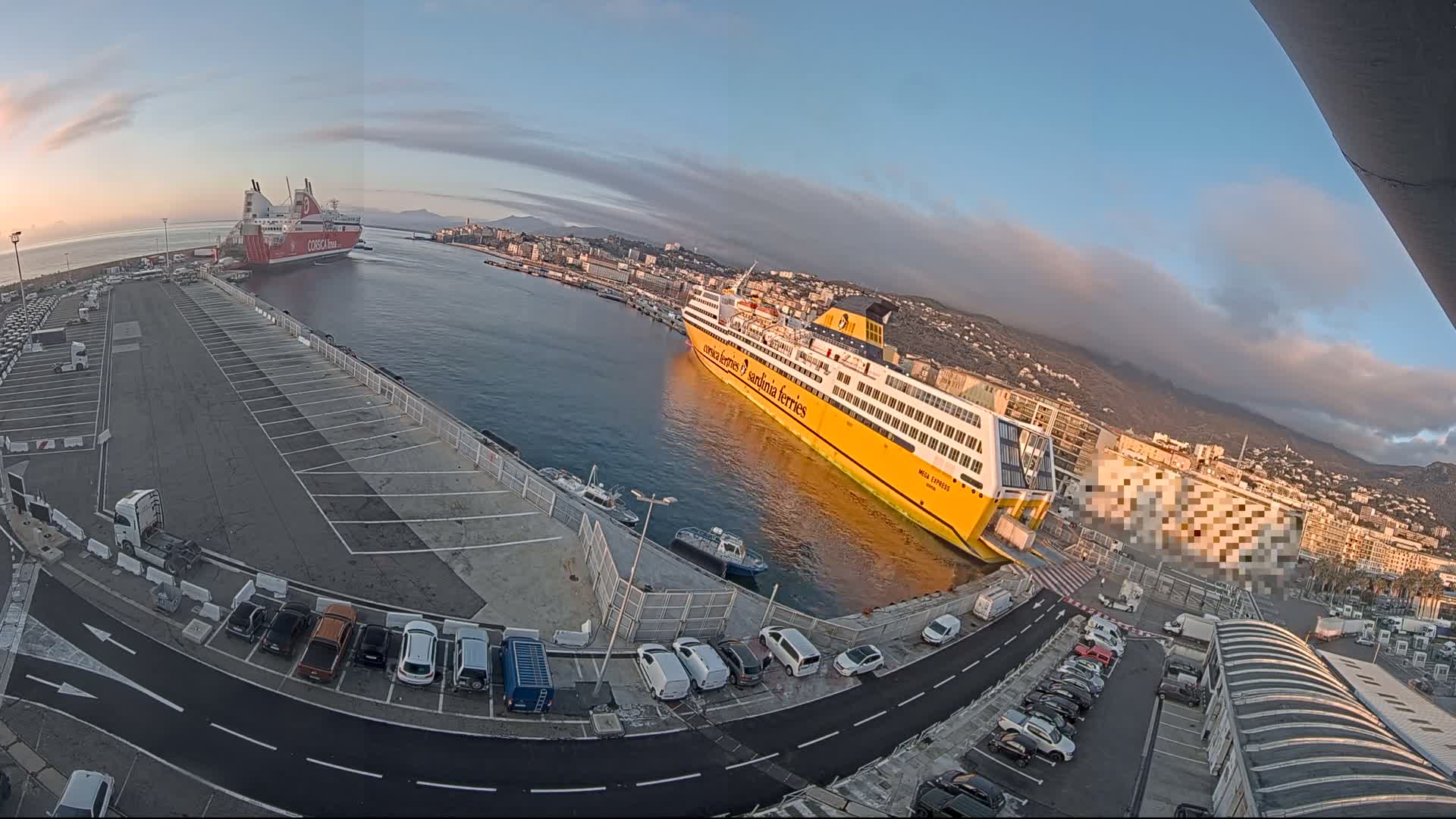 A high-angle, wide shot shows a port with two large ferries docked, one red and one yellow, under a mostly clear sky with some clouds at sunrise, and cars parked in a lot nearby.