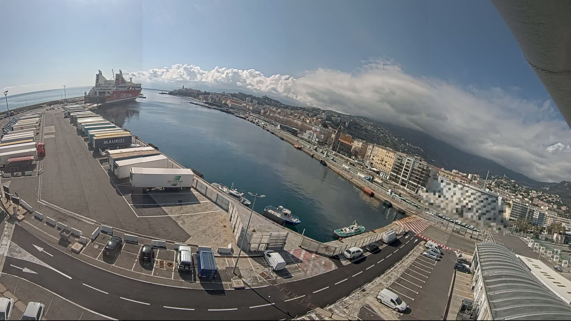 A ferry is docked at a harbor, numerous trucks are parked in a lot, and a city is visible in the background under a partly cloudy sky.