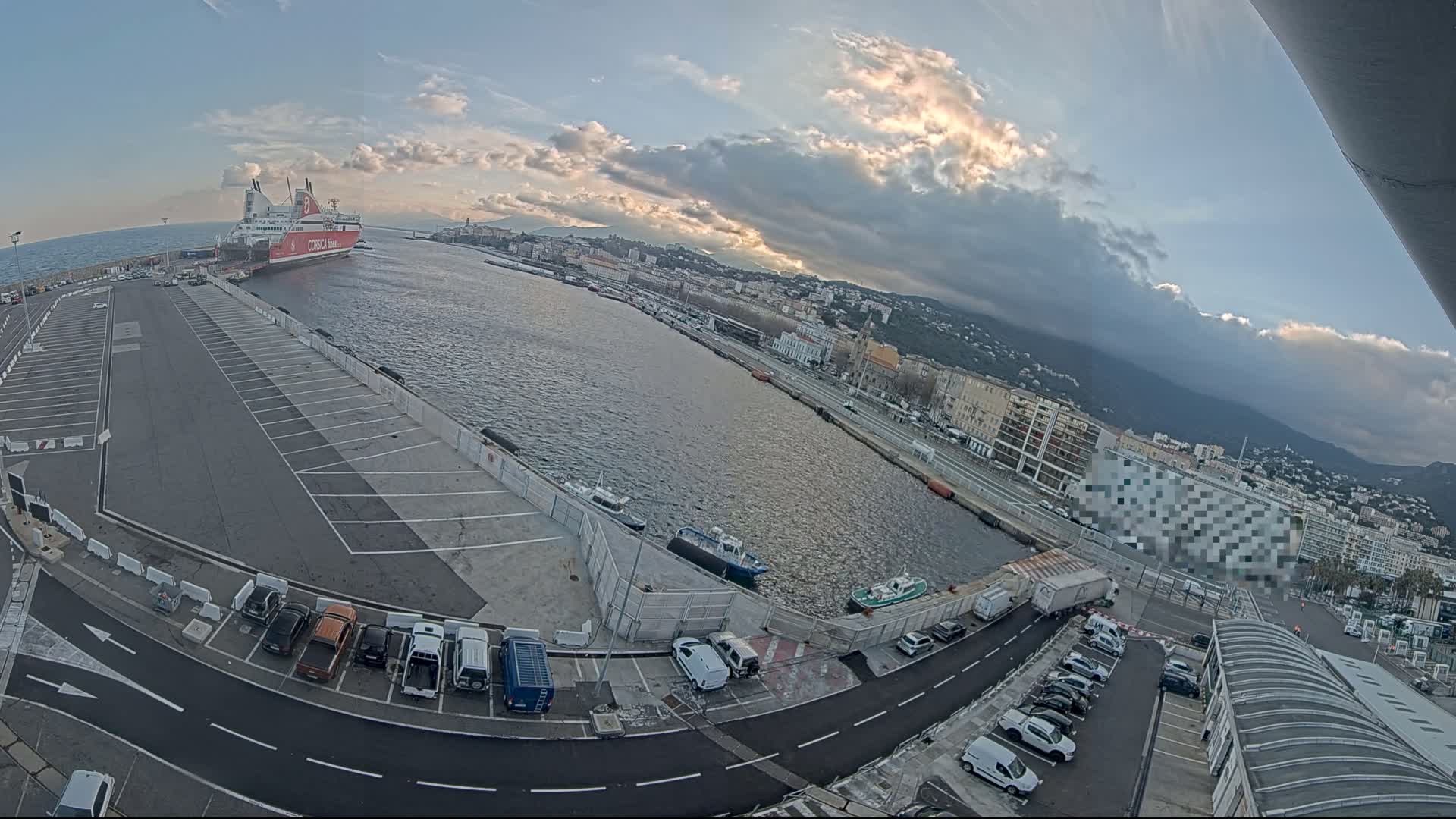 A large ferry is docked at a harbor, with several smaller boats and cars parked in the surrounding parking lots under partly cloudy skies.