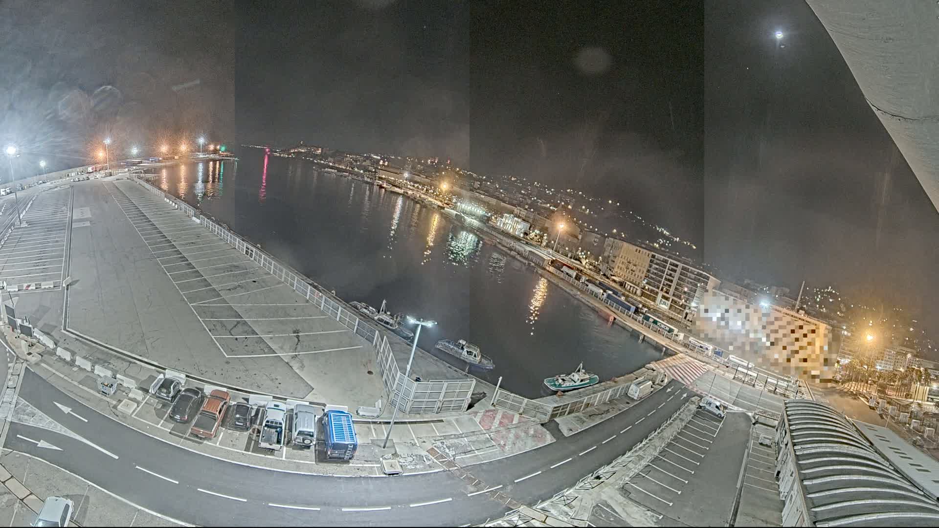 A nighttime aerial view of a harbor with a mostly empty parking lot, several small boats moored in the water, and a city illuminated in the background under a clear night sky.