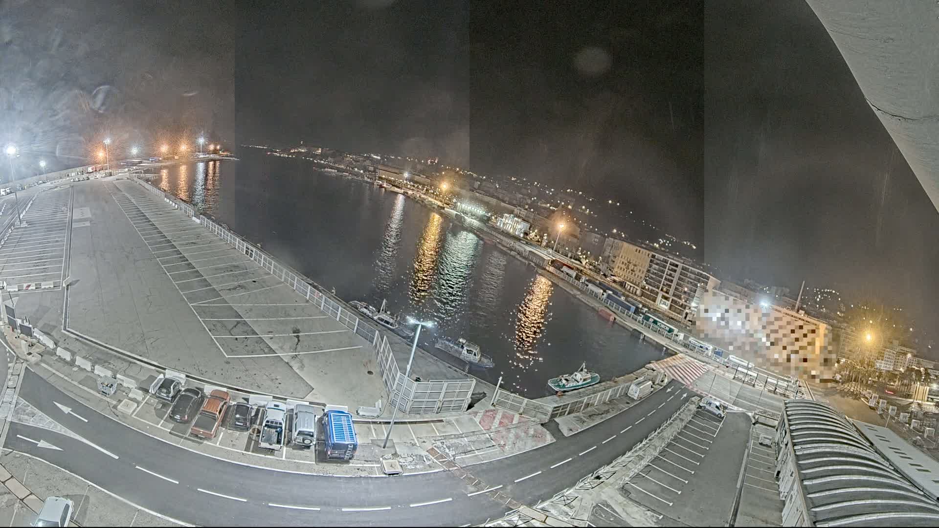 An empty parking lot at night next to a calm body of water with boats and a city lit up in the background.