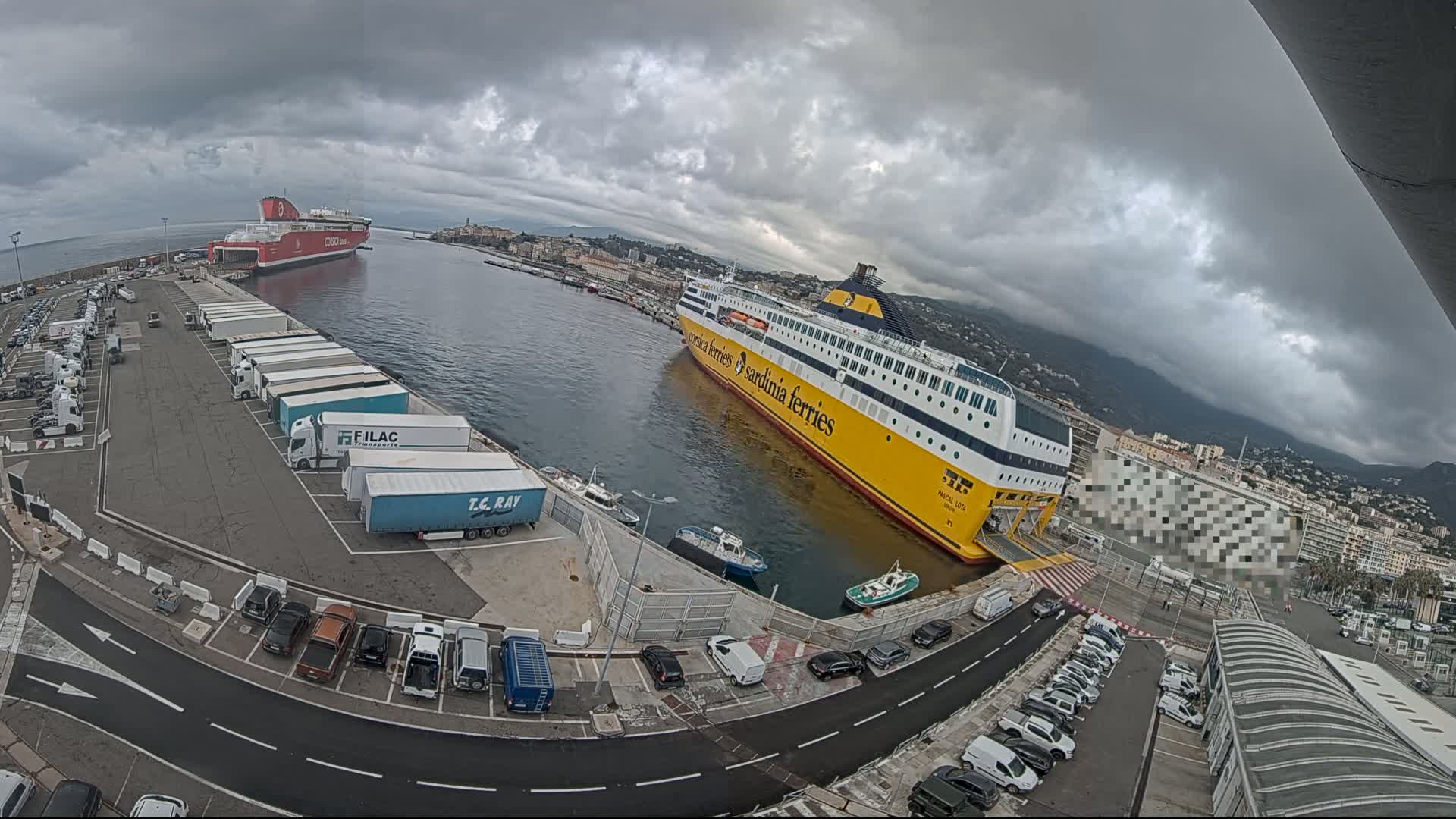Here is a description of the image in a single sentence: A large ferry is docked at a port under a cloudy sky, with another ferry visible in the distance, numerous cars and trucks parked nearby.
