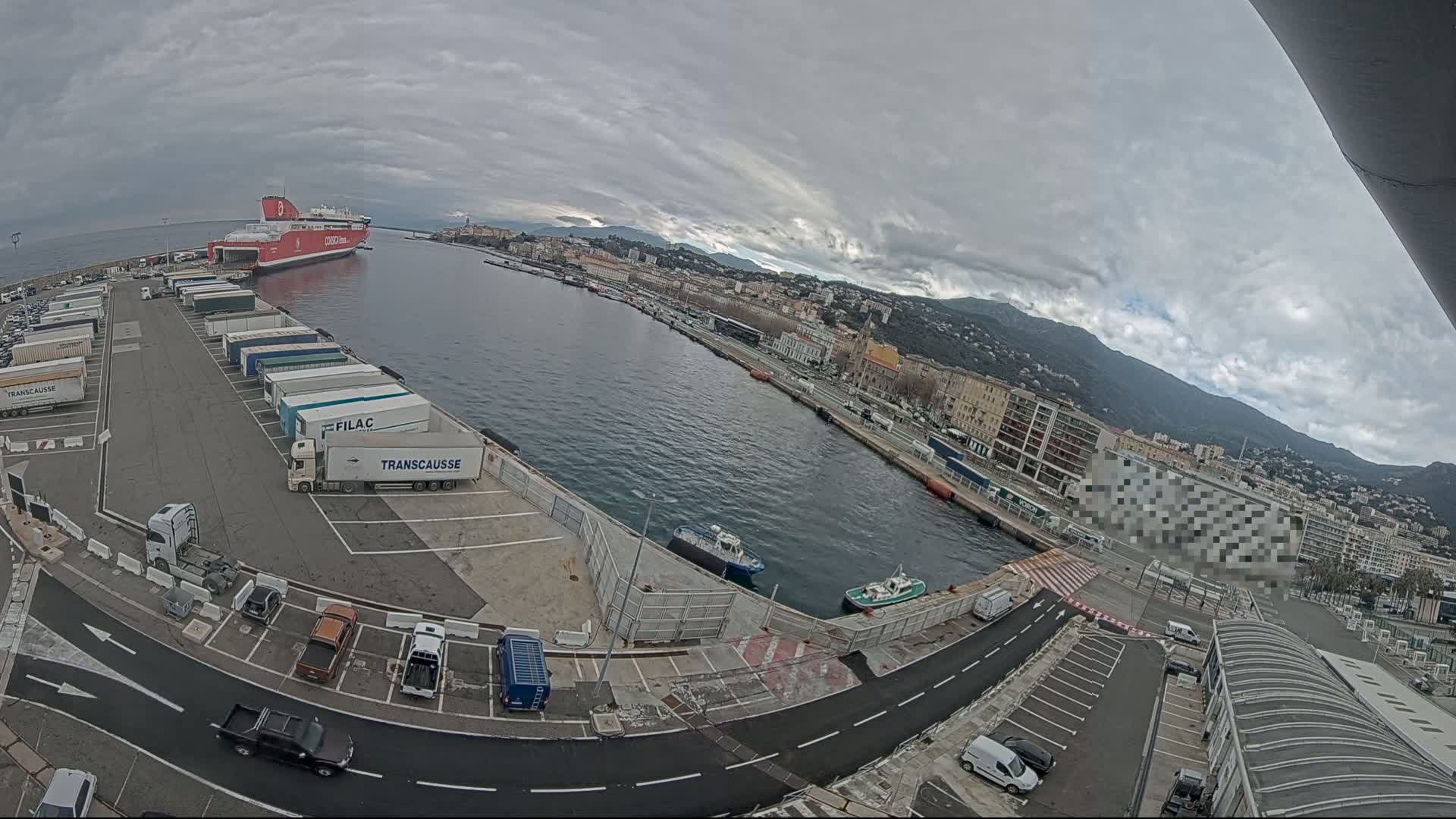 A red ferry is docked at a port in a city under an overcast sky, with numerous trucks and cars parked nearby and smaller boats in the water.
