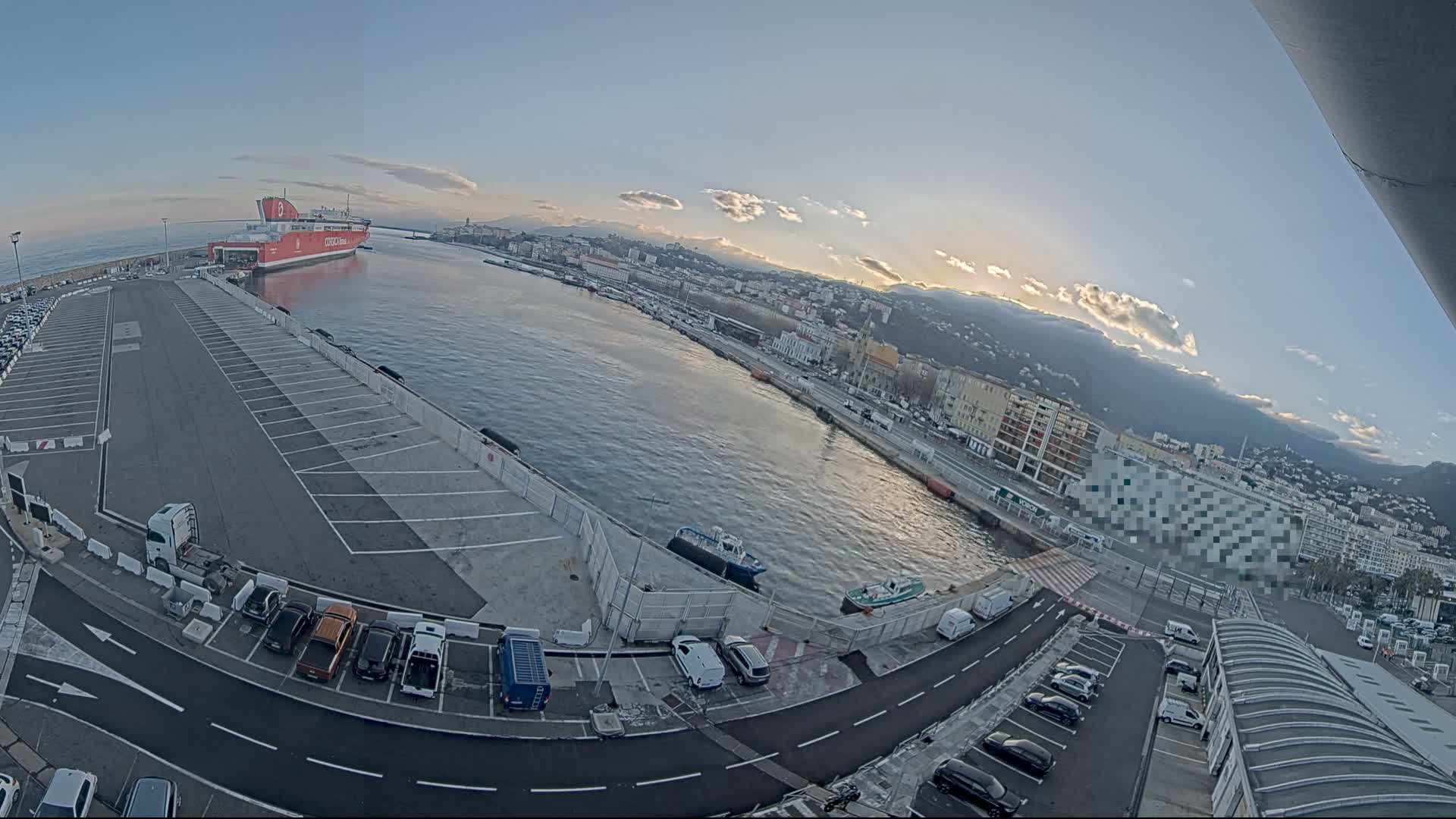 A large red ferry is docked at a port city under a partly cloudy sunset sky, with a parking lot and several smaller vessels visible.