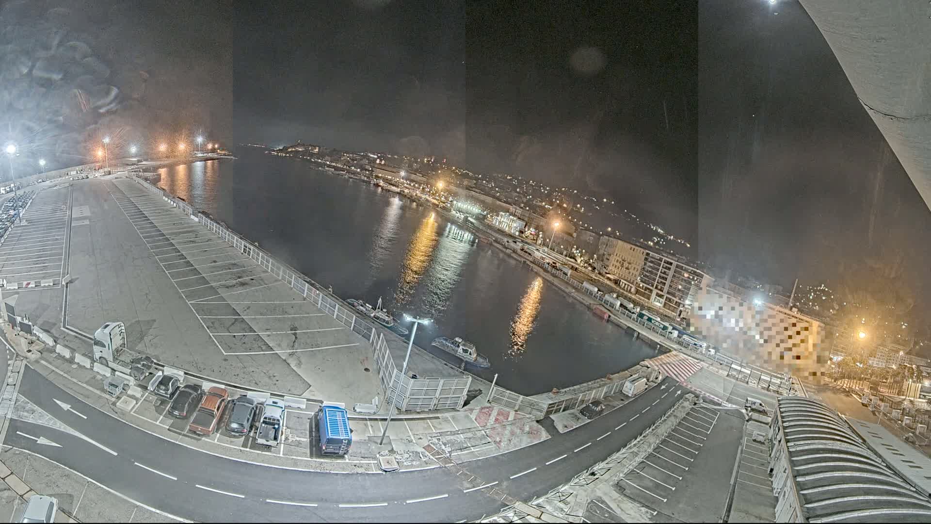 An empty parking lot at night next to a calm body of water with docked boats and a city lit up in the background.