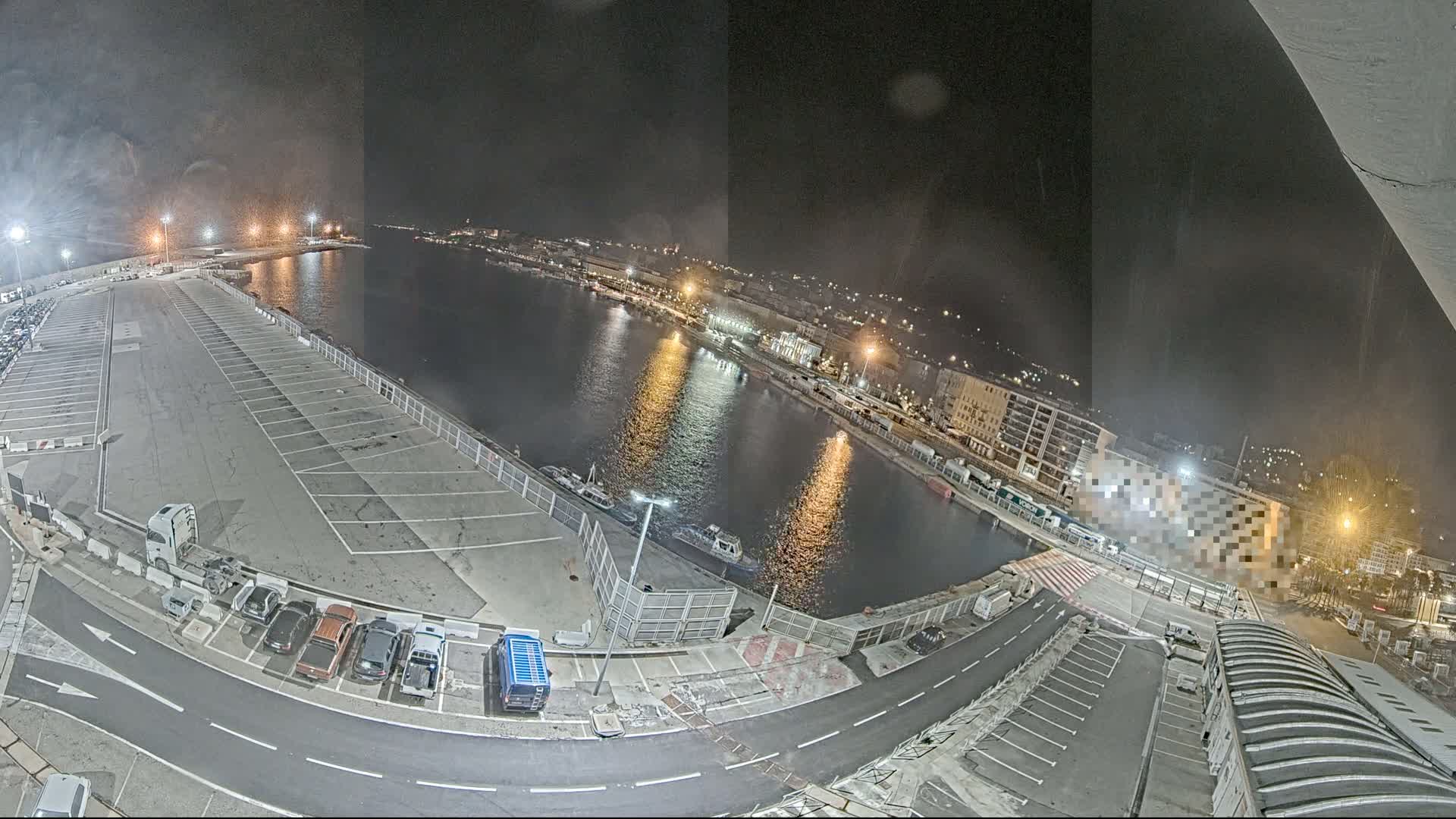 A nighttime aerial view shows a harbor with a parking lot, several vehicles, and a few small boats on the water.