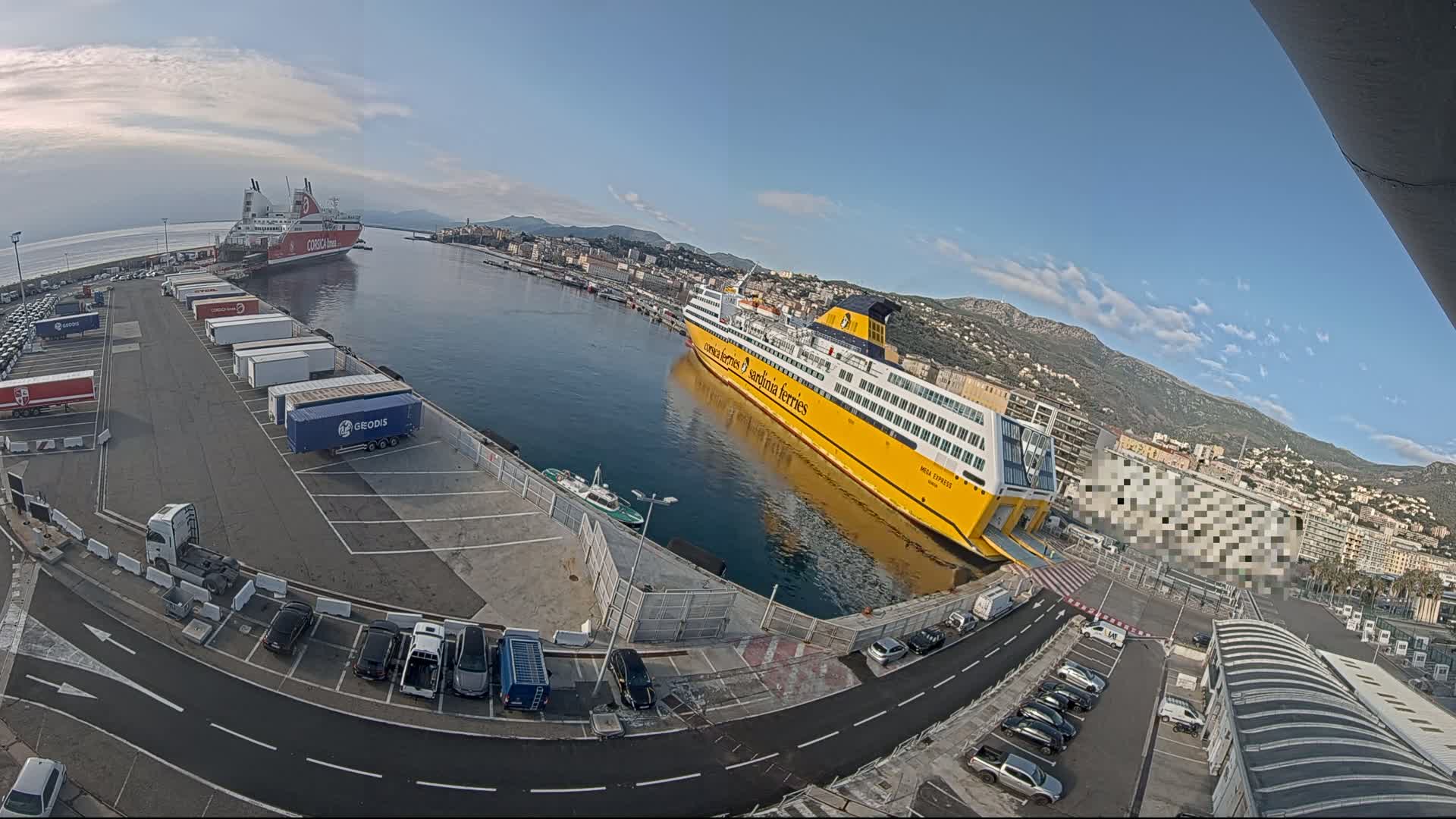 A harbor with two large ferries docked, numerous cars and trucks parked in a lot, and a city and mountains in the background under a mostly clear sky.