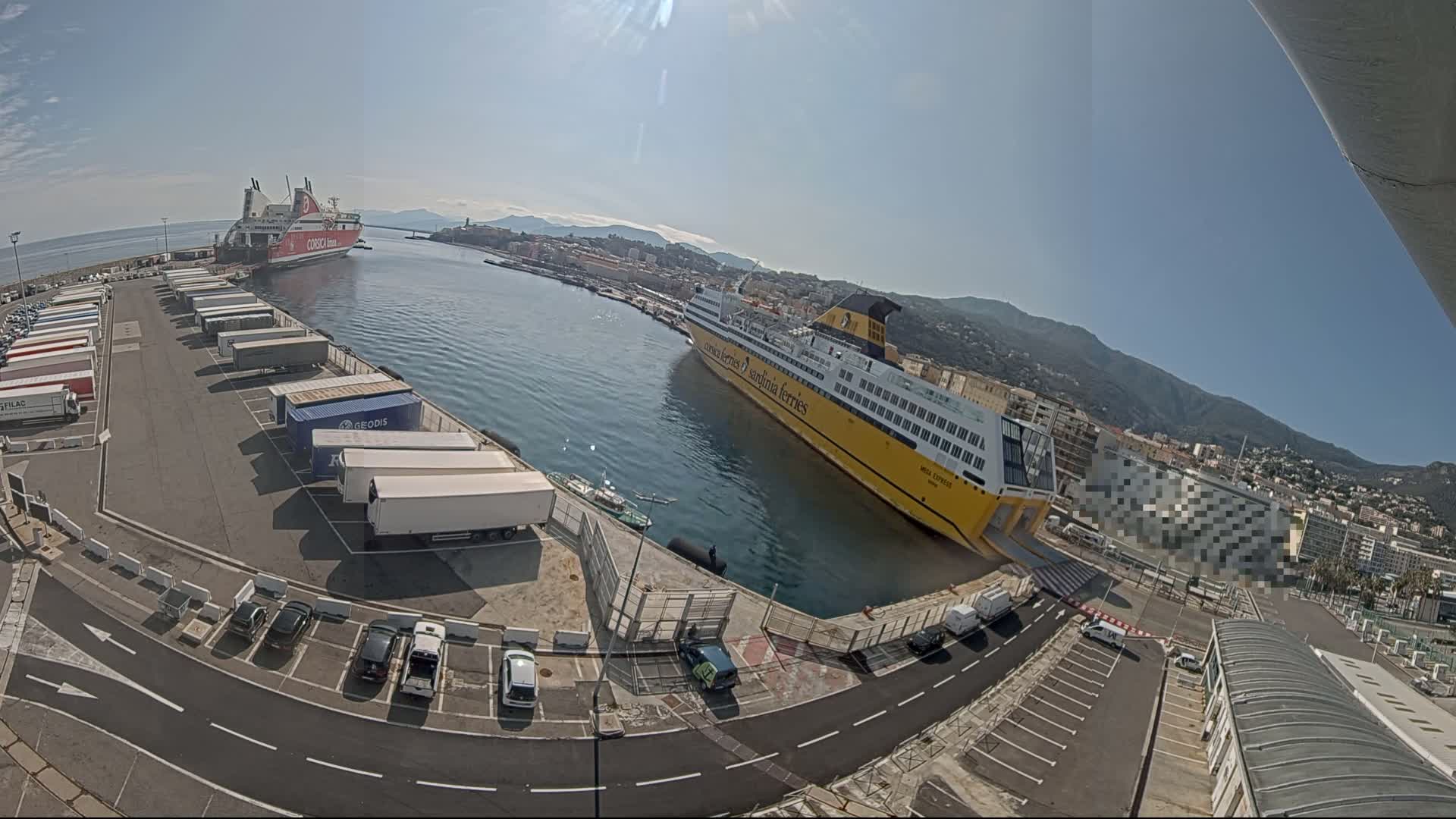 A large yellow ferry is docked at a port with a smaller red ferry visible in the distance under a sunny sky, numerous trucks and cars are parked in the port area.