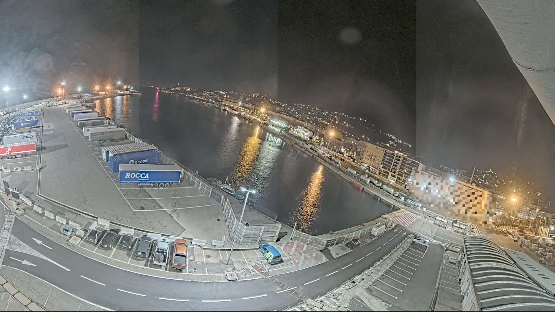 A nighttime view of a harbor at a port, showing a calm body of water reflecting city lights, alongside a parking lot filled with trucks and cars.
