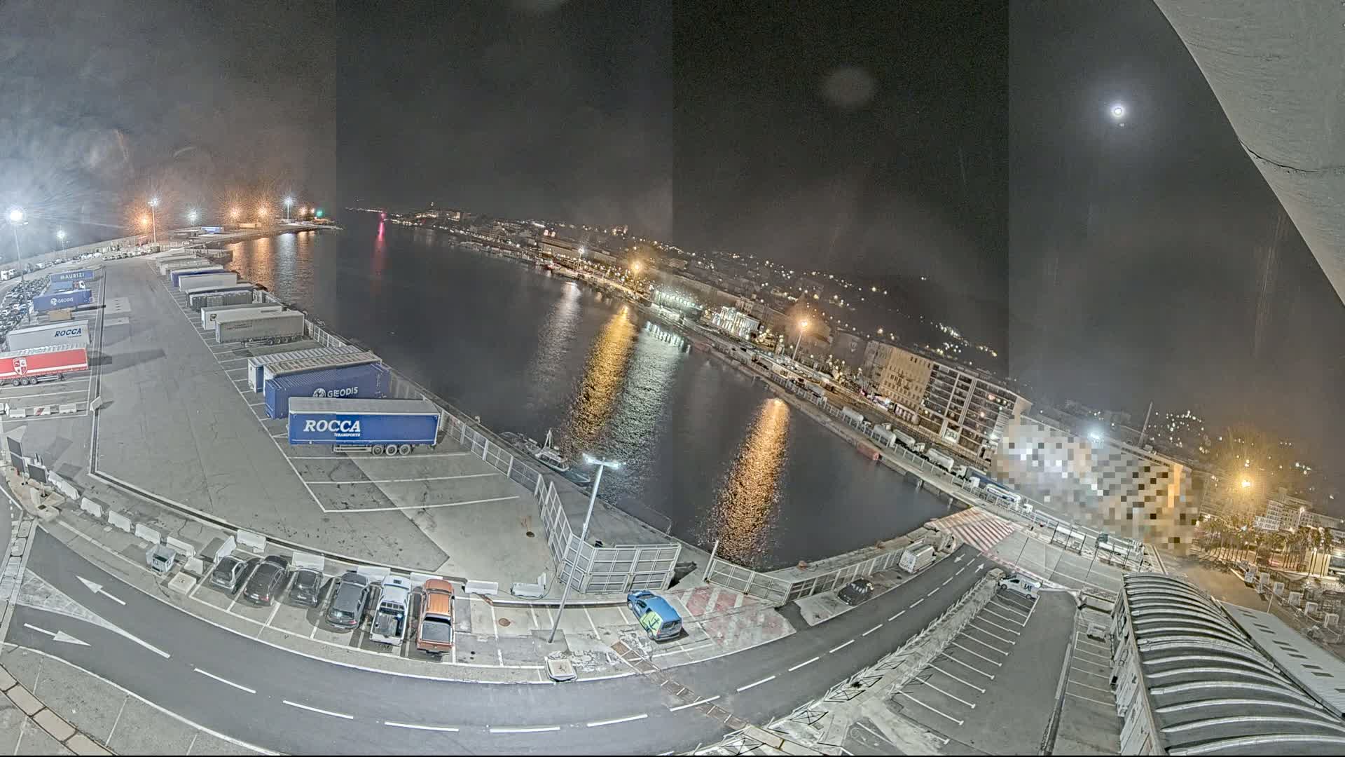 A nighttime, high-angle view of a harbor with a moon in the sky, showing a parking lot filled with trucks and cars, a calm body of water reflecting city lights, and buildings along the waterfront.