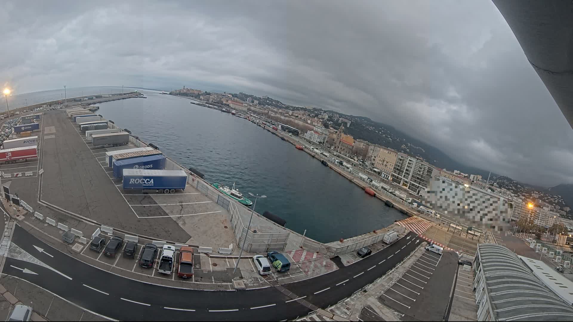 A harbor with overcast skies, featuring numerous parked trucks and cars, a body of water with a small vessel, and a city in the background.