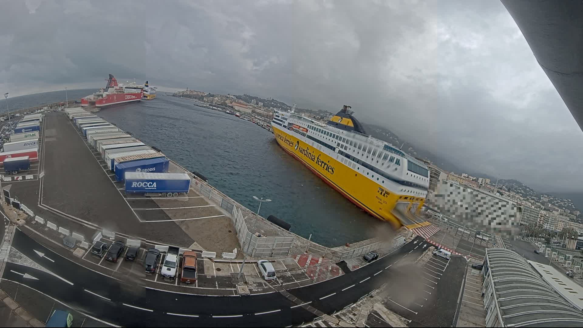 A large yellow ferry is docked at a port, with a smaller red ferry further in the background, under an overcast sky.