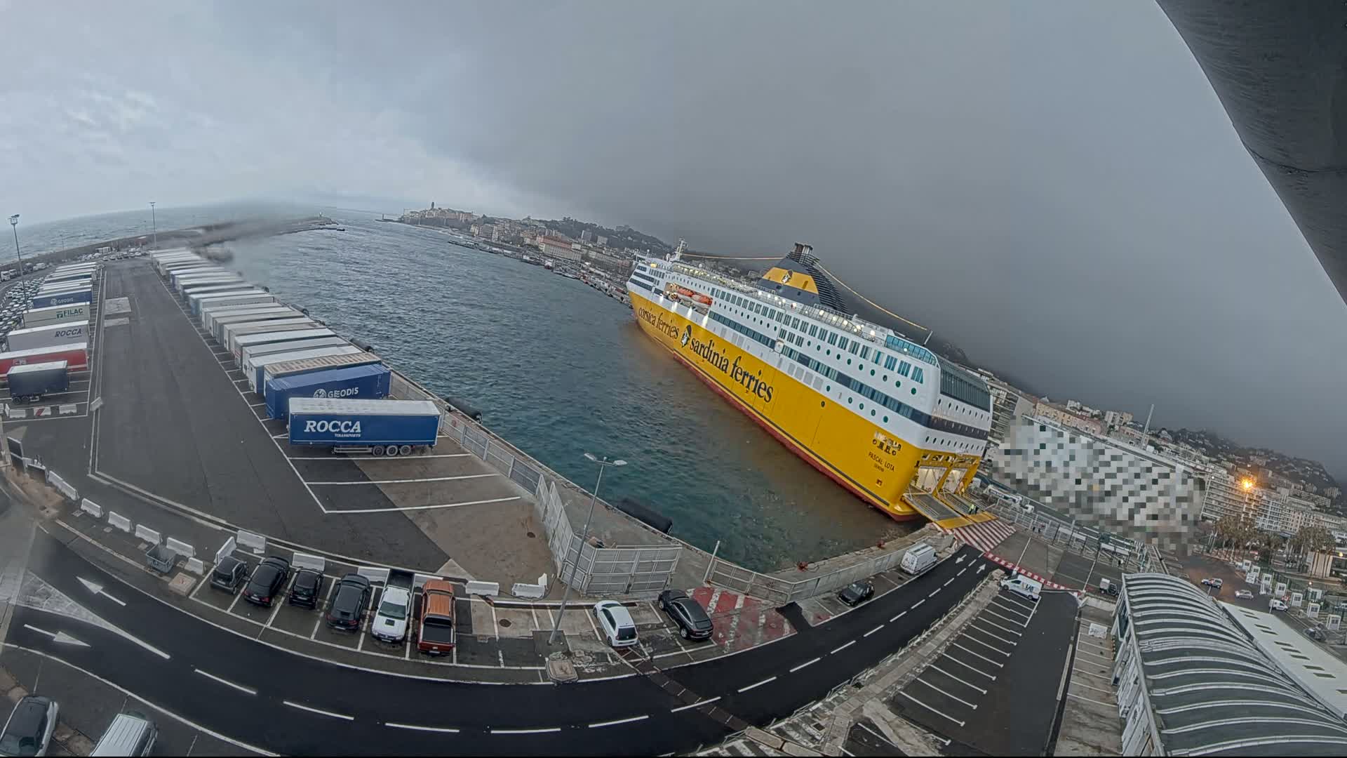 A large yellow ferry is docked at a port under an overcast sky, with numerous trucks and cars parked nearby.