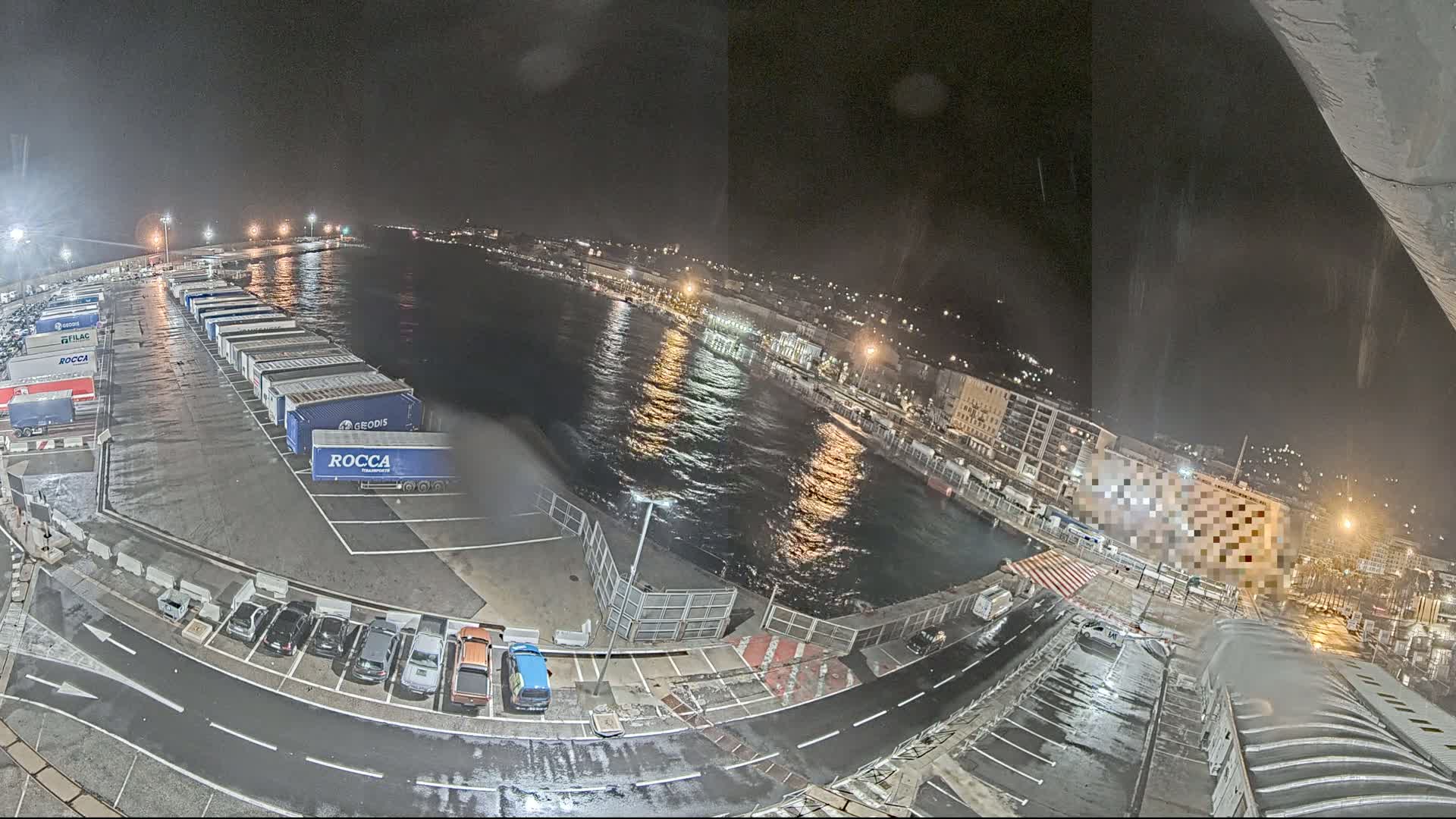 A nighttime view of a harbor with numerous semi-trailer trucks parked in a lot, cars in a nearby parking area, and choppy water in the harbor under a dark, possibly rainy sky.