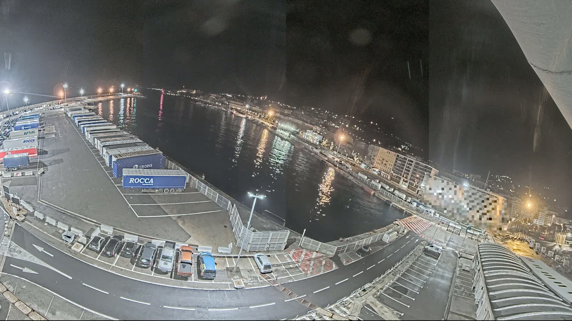 A nighttime view of a harbor with numerous parked trucks and cars, a calm body of water reflecting city lights, and some rain.