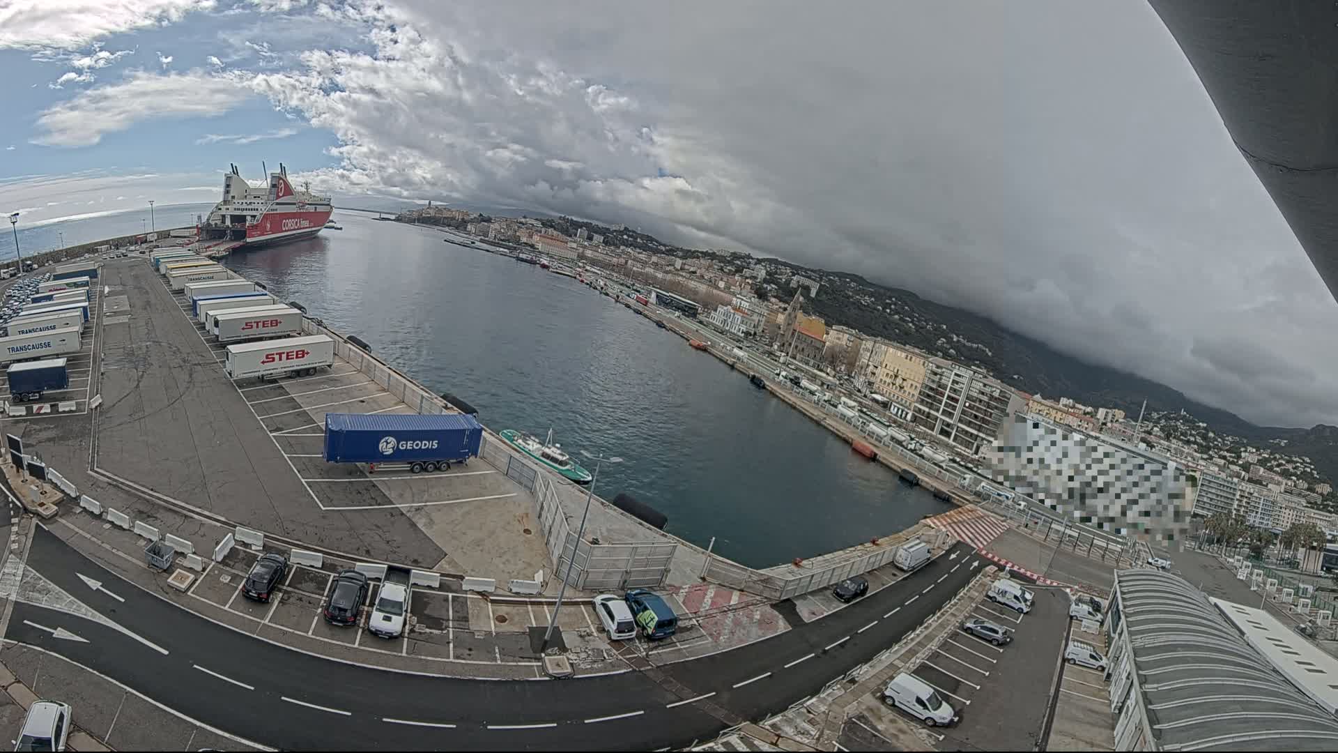 A ferry is docked at a port on an overcast day, with numerous trucks and cars parked in the surrounding area.