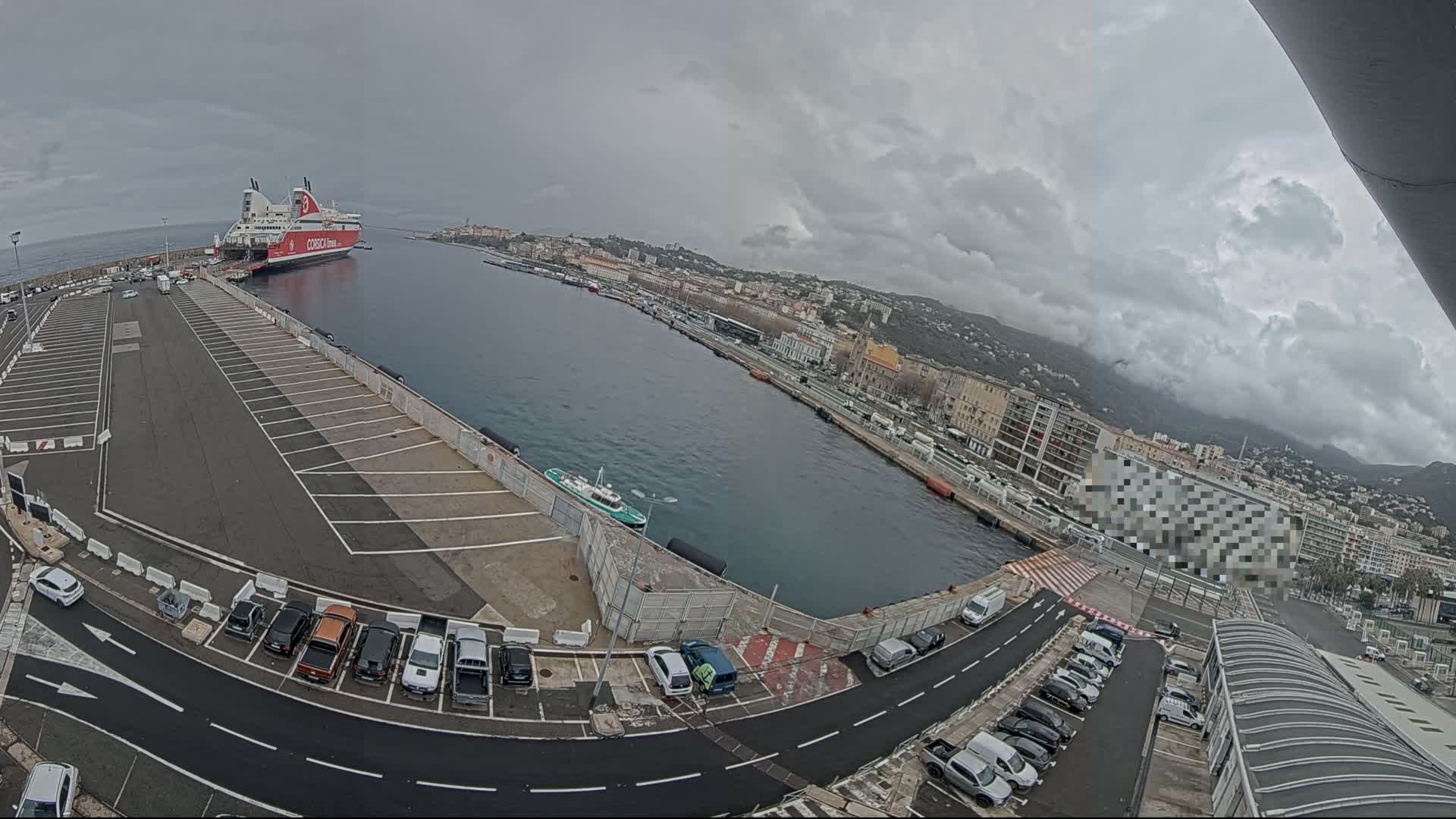 A large ferry is docked at a port in a city on a cloudy day, with several cars parked in lots surrounding the harbor.