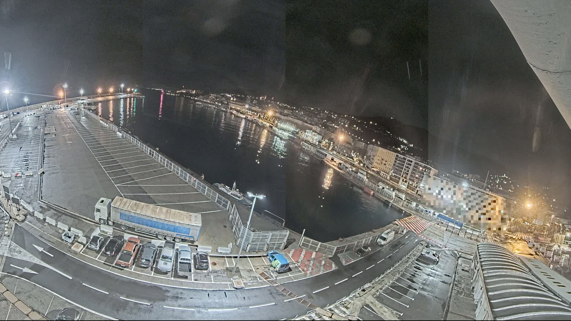 A nighttime aerial view of a harbor with a mostly empty parking lot, several vehicles, and a city illuminated across the water.