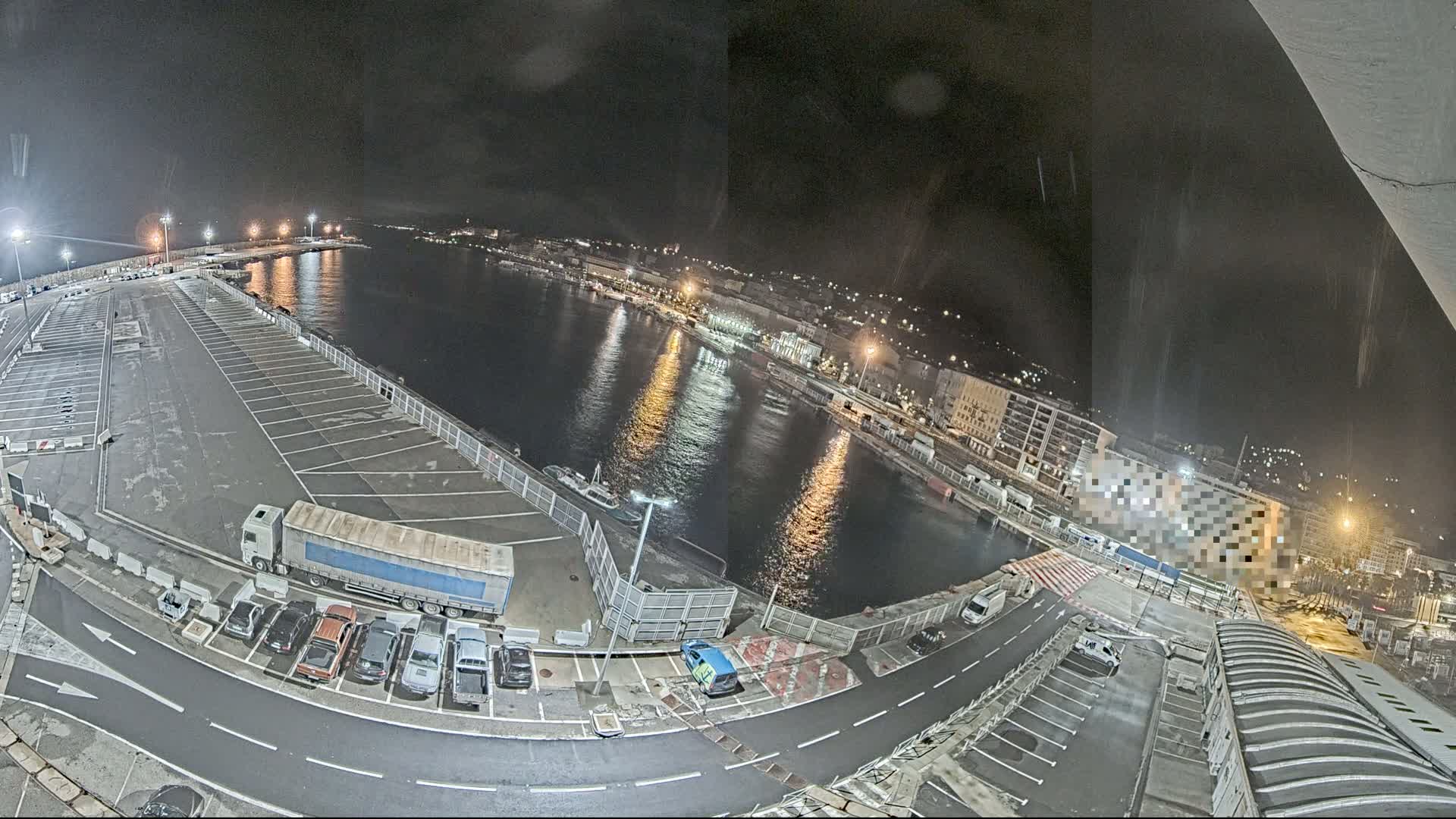 A nighttime, high-angle view of a harbor with a large empty parking lot next to a calm body of water,  surrounded by buildings with lights.
