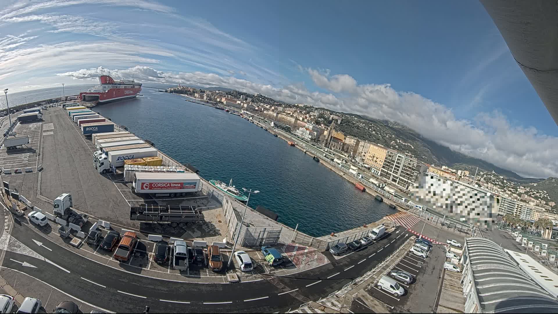 A ferry is docked at a harbor on a partly cloudy day, with numerous trucks and cars parked in the surrounding lots and a city visible in the background.