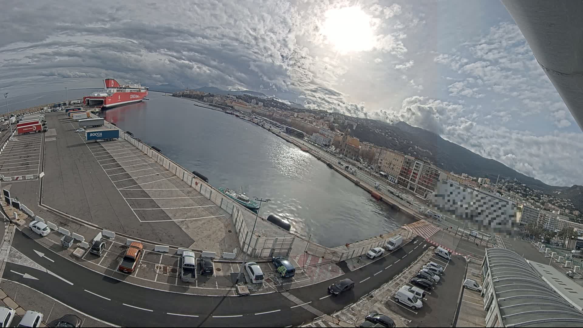 A large red ferry is docked at a port, with a city and mountains visible in the background under a partly cloudy sky.