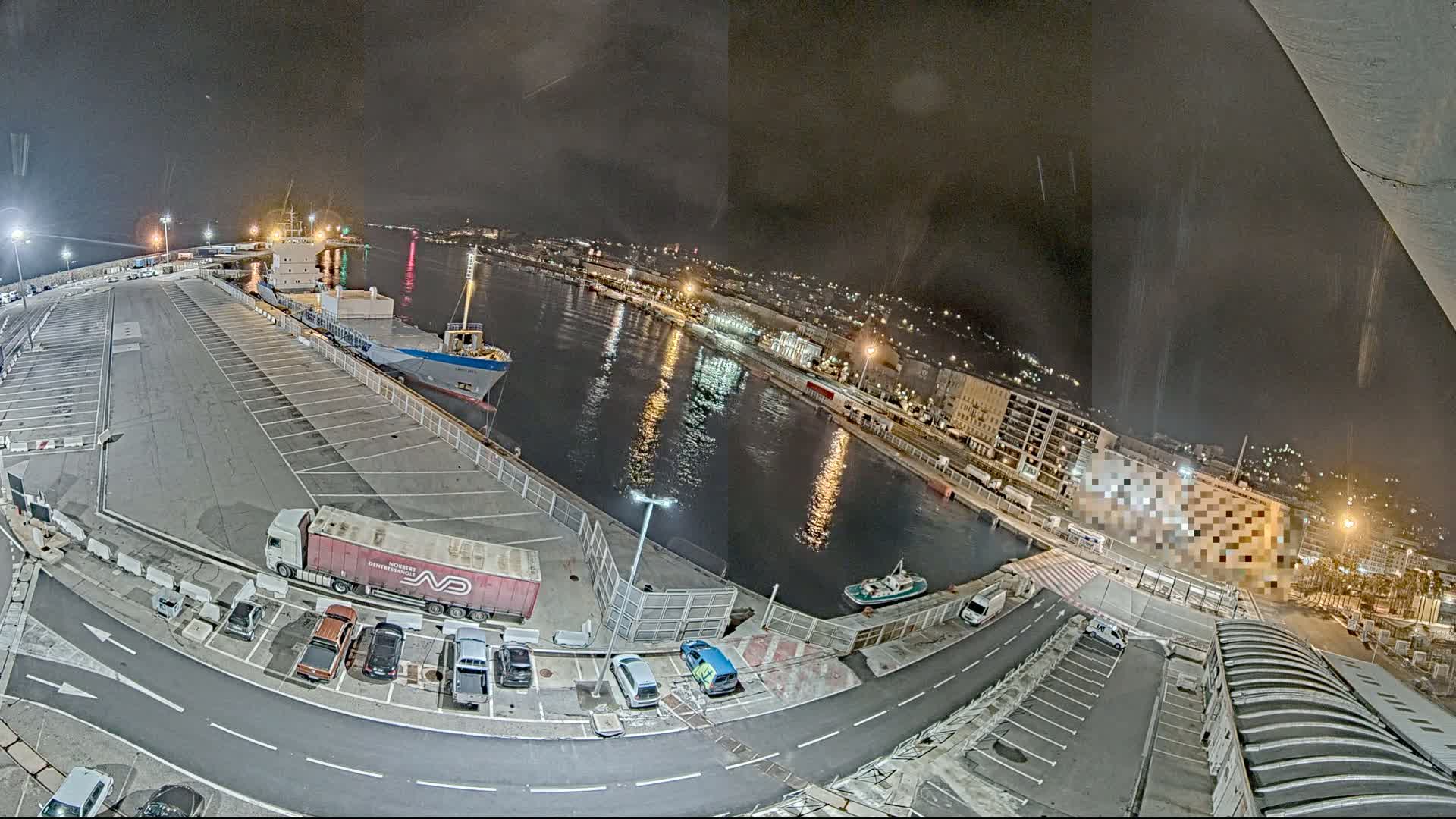 A nighttime high-angle view of a harbor with docked ships, a parking lot filled with cars and trucks, and a city illuminated in the background under a dark, possibly slightly rainy sky.