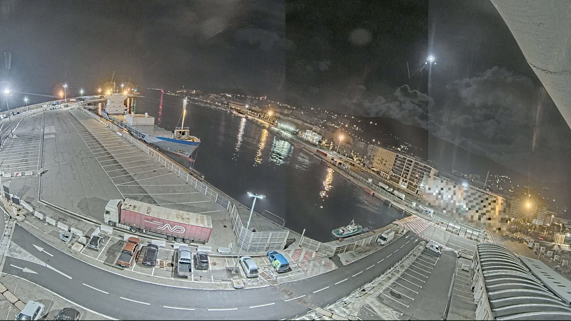 A nighttime, high-angle view of a harbor with a cargo ship docked, several vehicles parked in a lot, and a small boat on the water under a partly cloudy sky.