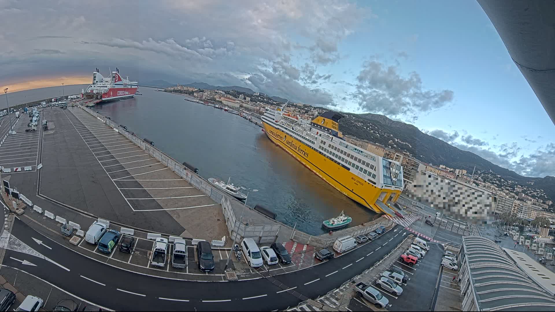 Two large ferries are docked at a port with a mostly cloudy sky, and cars are parked in a lot nearby, with mountains visible in the background.