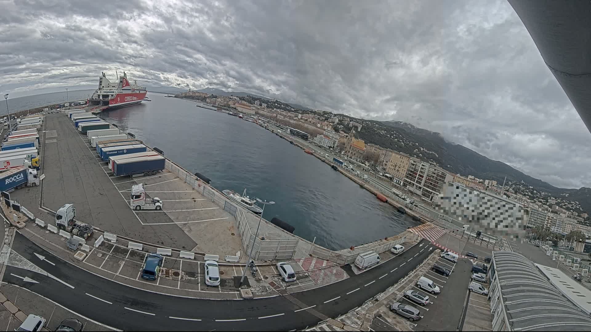 A large red and white ferry is docked at a harbor, with numerous trucks and cars parked in the surrounding lots under an overcast sky.