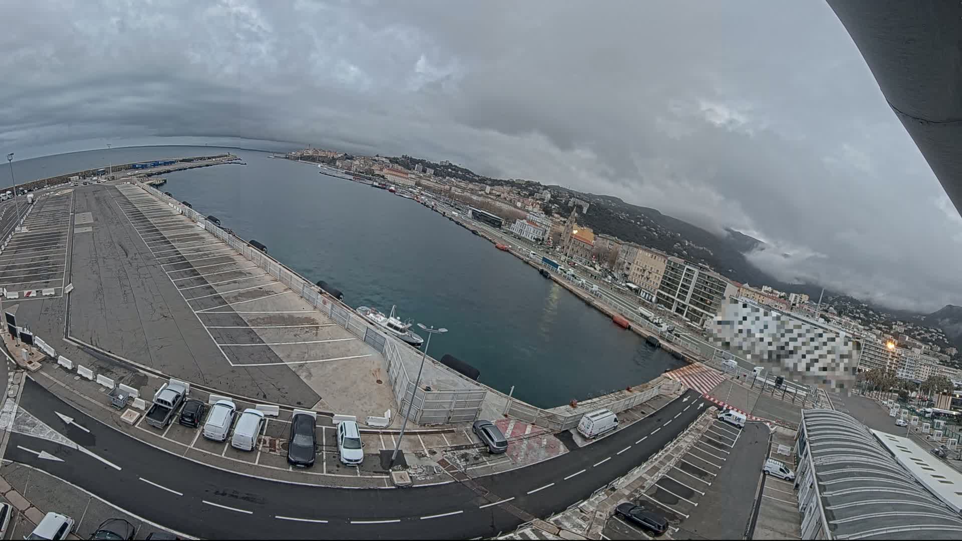 An overcast day shows a harbor with a large parking lot, several cars, and a city in the background, partially obscured by clouds covering the mountains.
