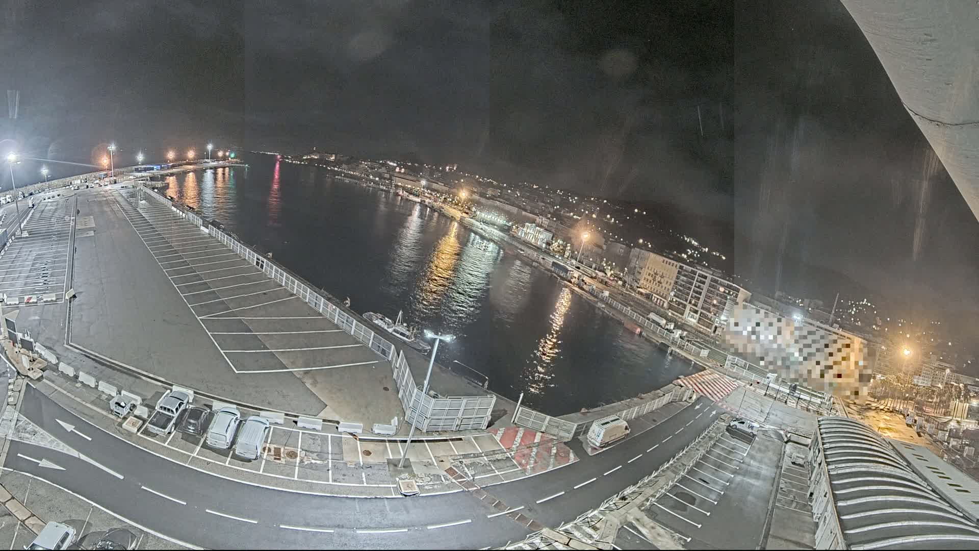A nighttime aerial view of a harbor with a parking lot, calm water reflecting lights from buildings on the shore, and a few vehicles parked near the water's edge.