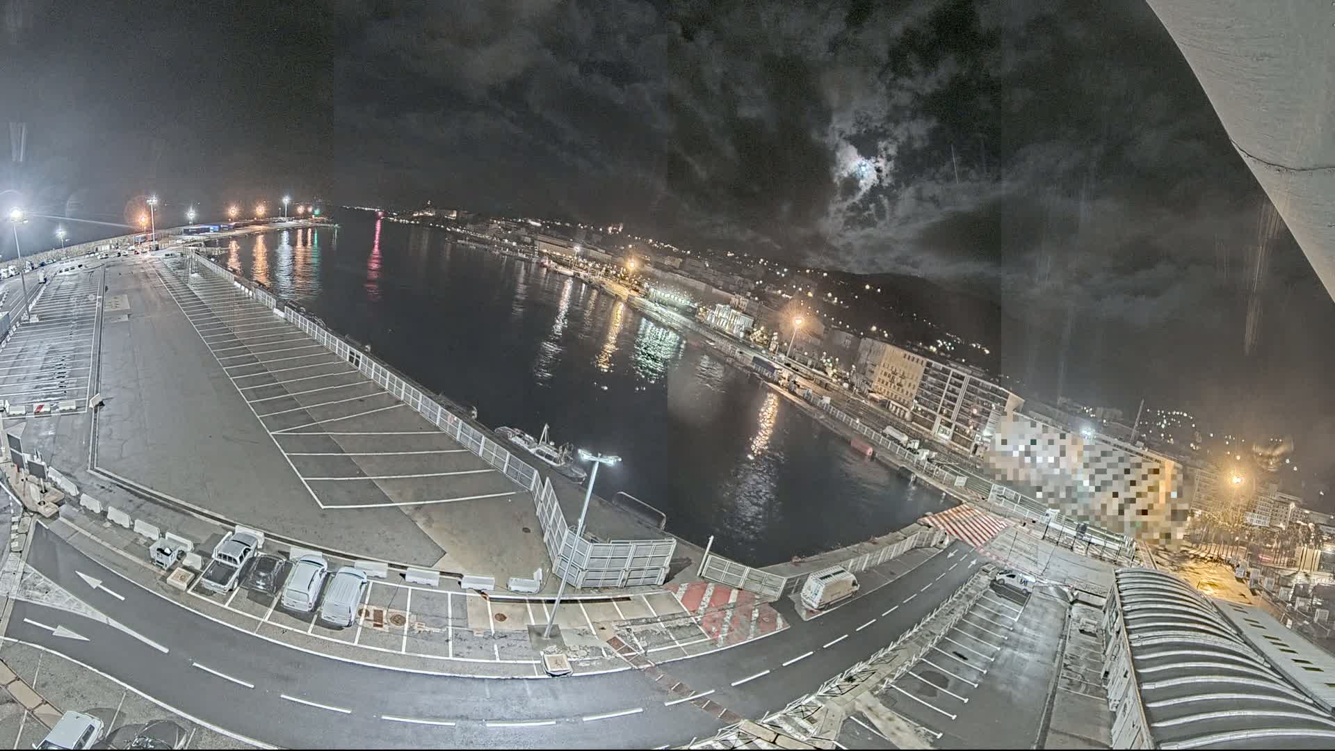 An empty parking lot at a harbor is illuminated at night under a partly cloudy sky, with a body of water reflecting city lights beyond a gated area.