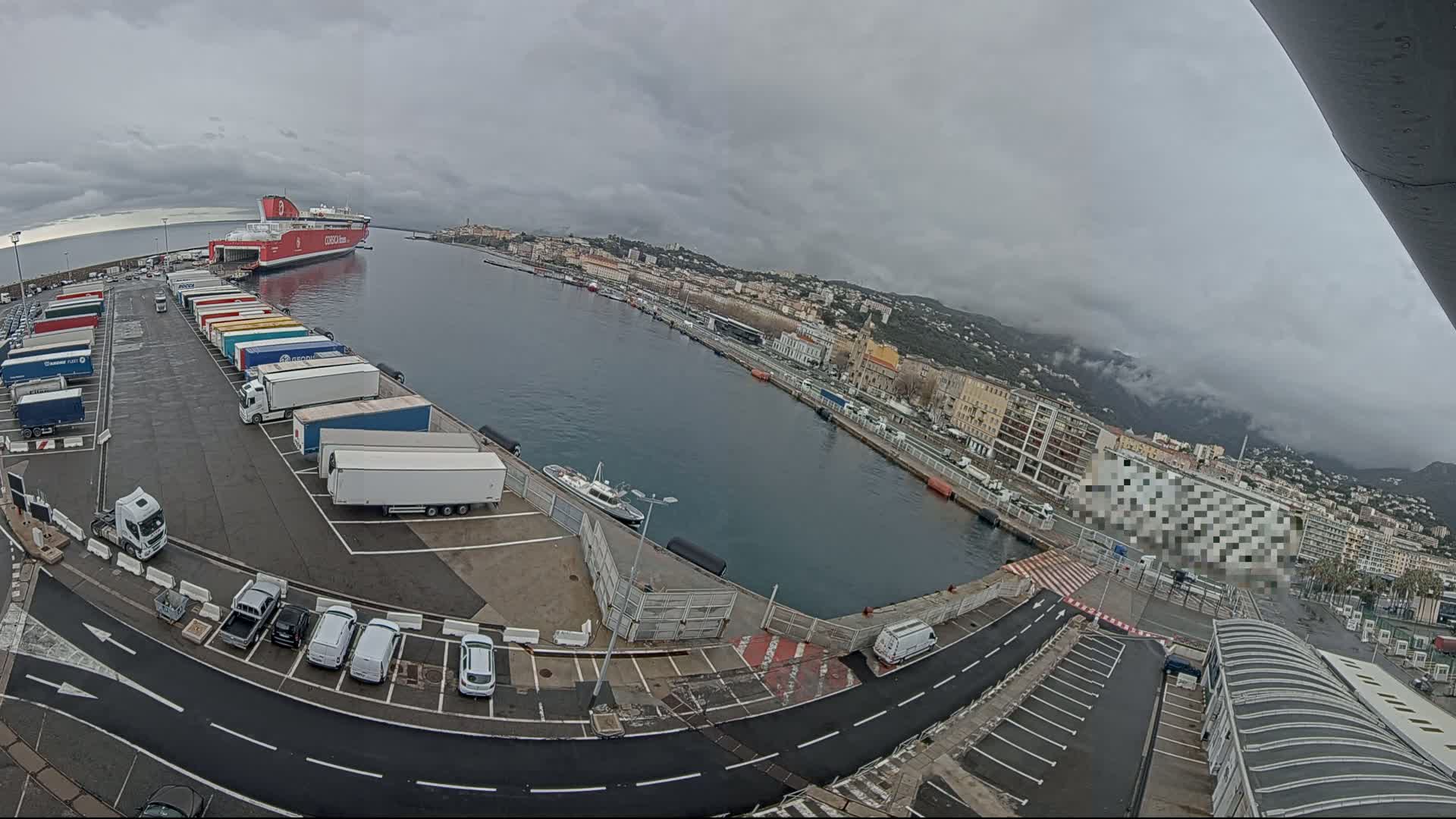 A large red ferry is docked at a port, with numerous trucks and cars parked in the surrounding area on an overcast day.