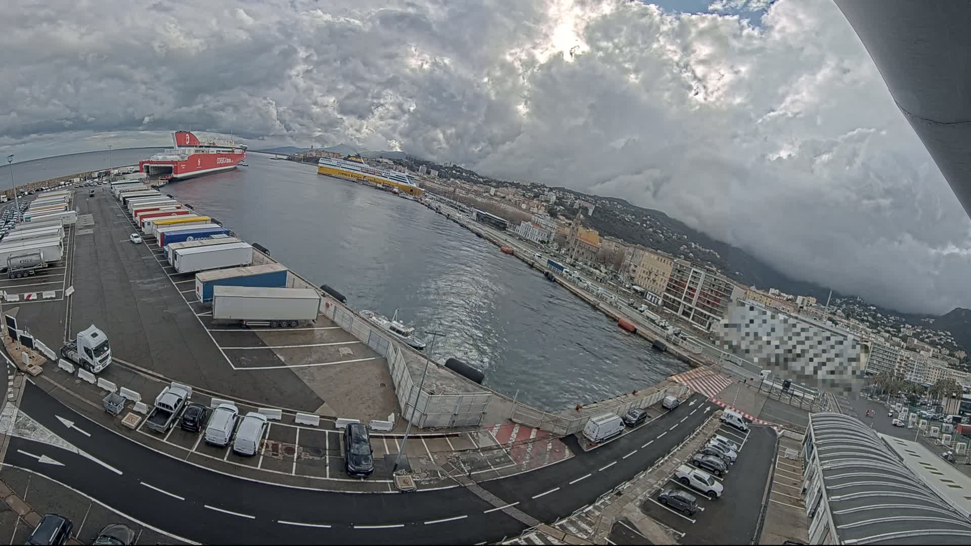 A ferry port on a cloudy day, with numerous trucks parked in the lot and several cars parked nearby, shows a large red ferry and a smaller yellow one in the harbor.