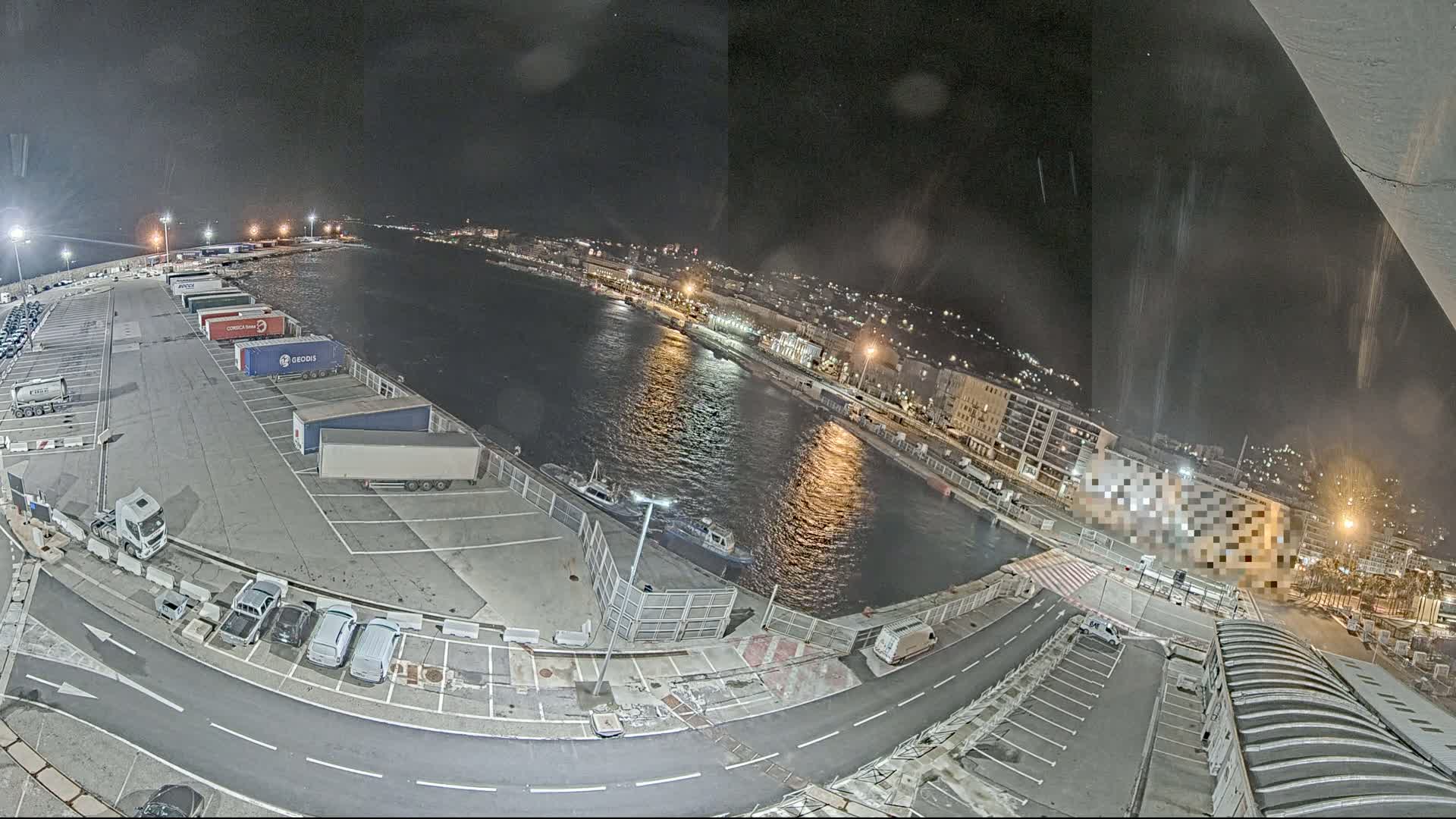 A nighttime aerial view of a harbor, showing a parking lot with numerous trucks and cars, a body of water with several boats, and a city skyline in the background under a dark, possibly slightly overcast sky.