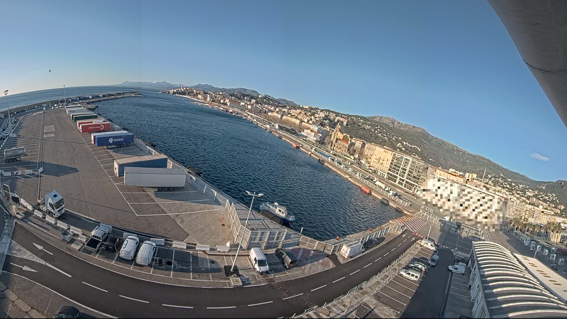 A high-angle, wide view of a harbor on a clear, sunny day shows a parking lot filled with semi-trucks and cars next to a calm body of water with a city and mountains in the background.
