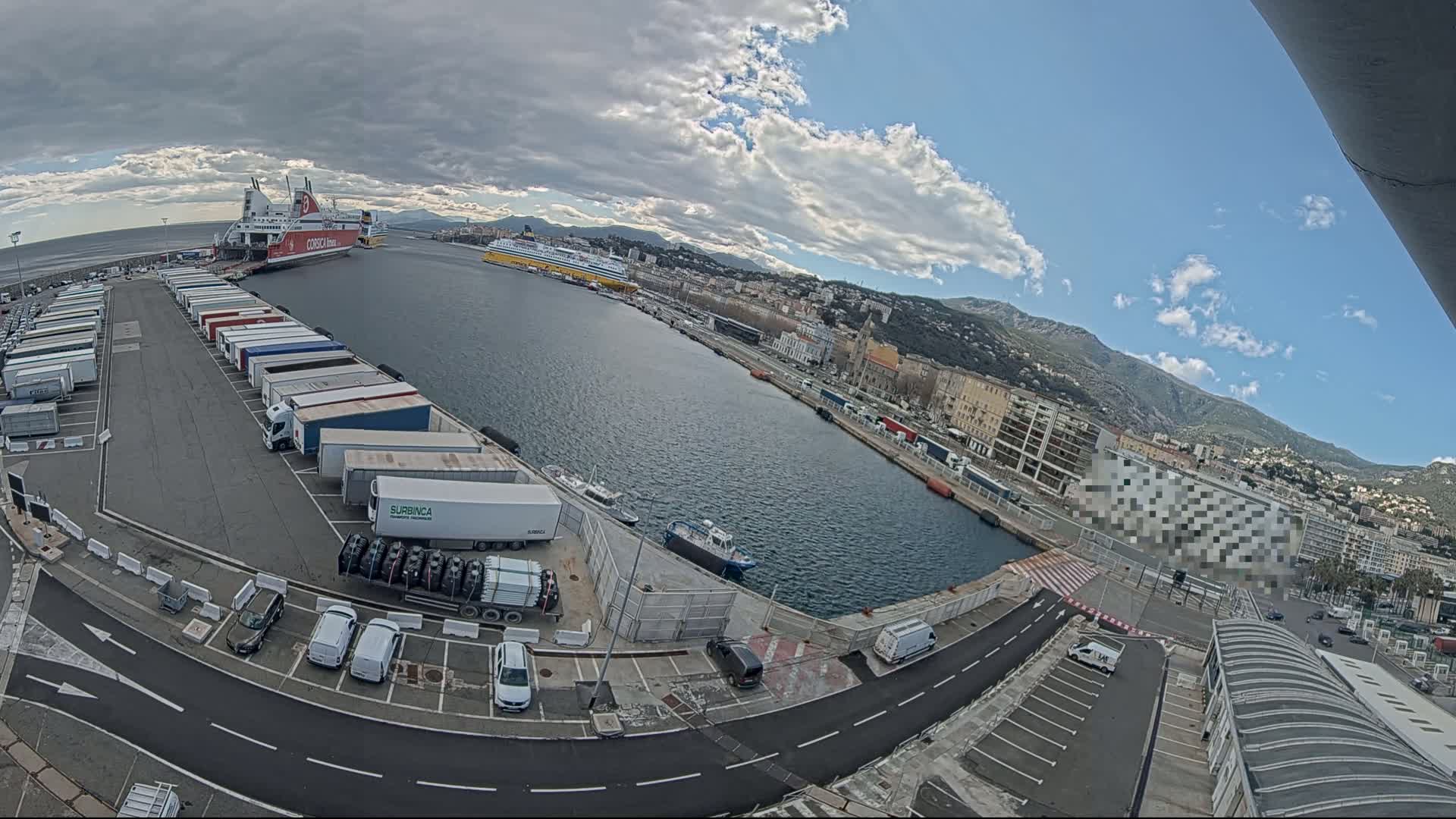 A partly cloudy day at a port shows docked ferries, numerous trucks and cars in parking lots, and a city with mountains in the background.