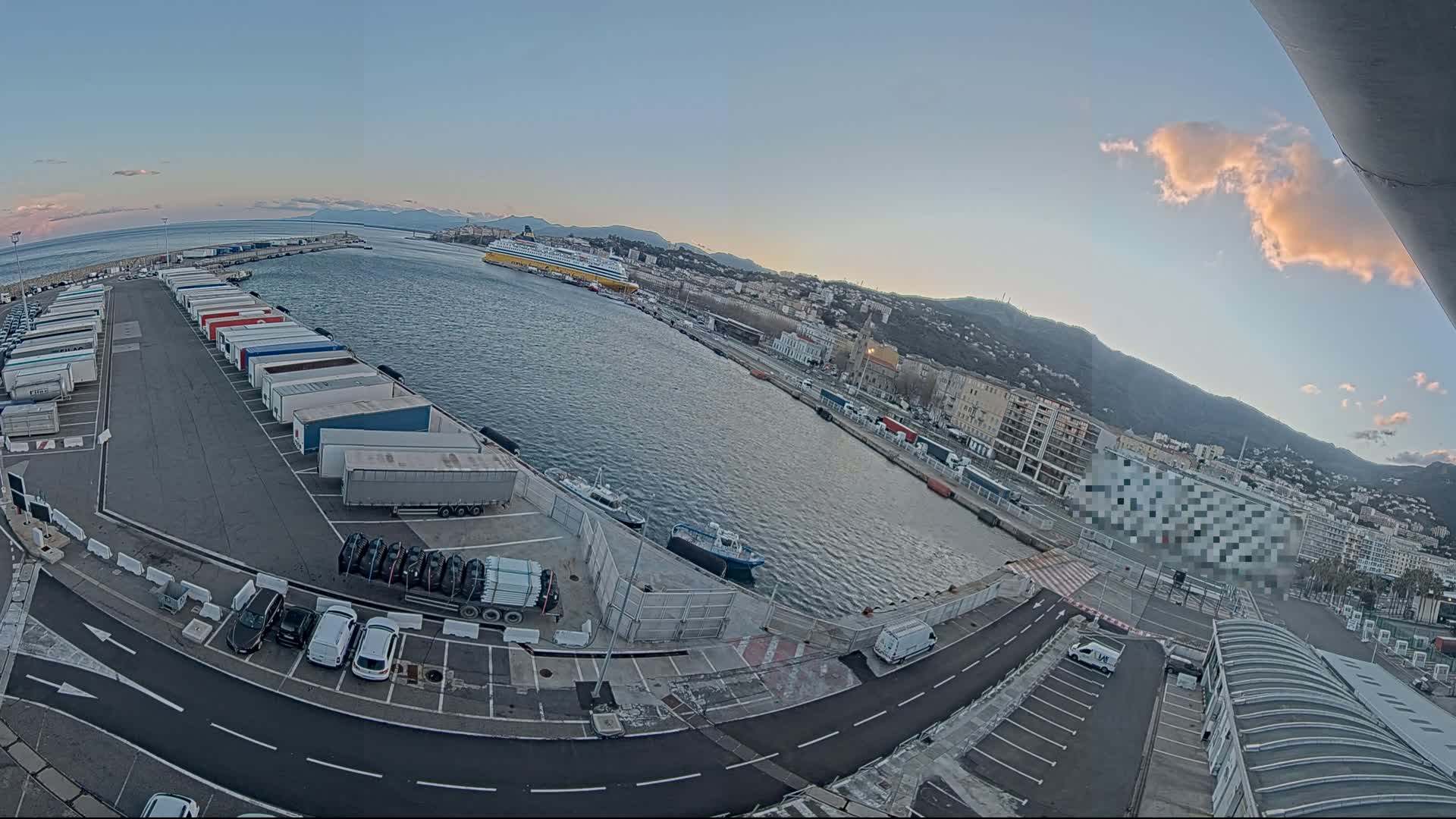 A harbor at sunset with numerous trucks and cars parked in a lot, a large ferry in the water, and a city and mountains in the background under a mostly clear sky with a few clouds.